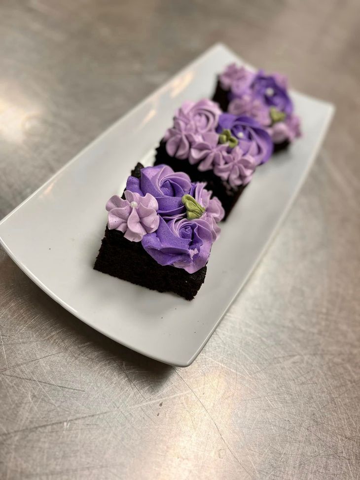 Three square chocolate brownies decorated with purple and lavender buttercream floral swirls on a white rectangular plate.