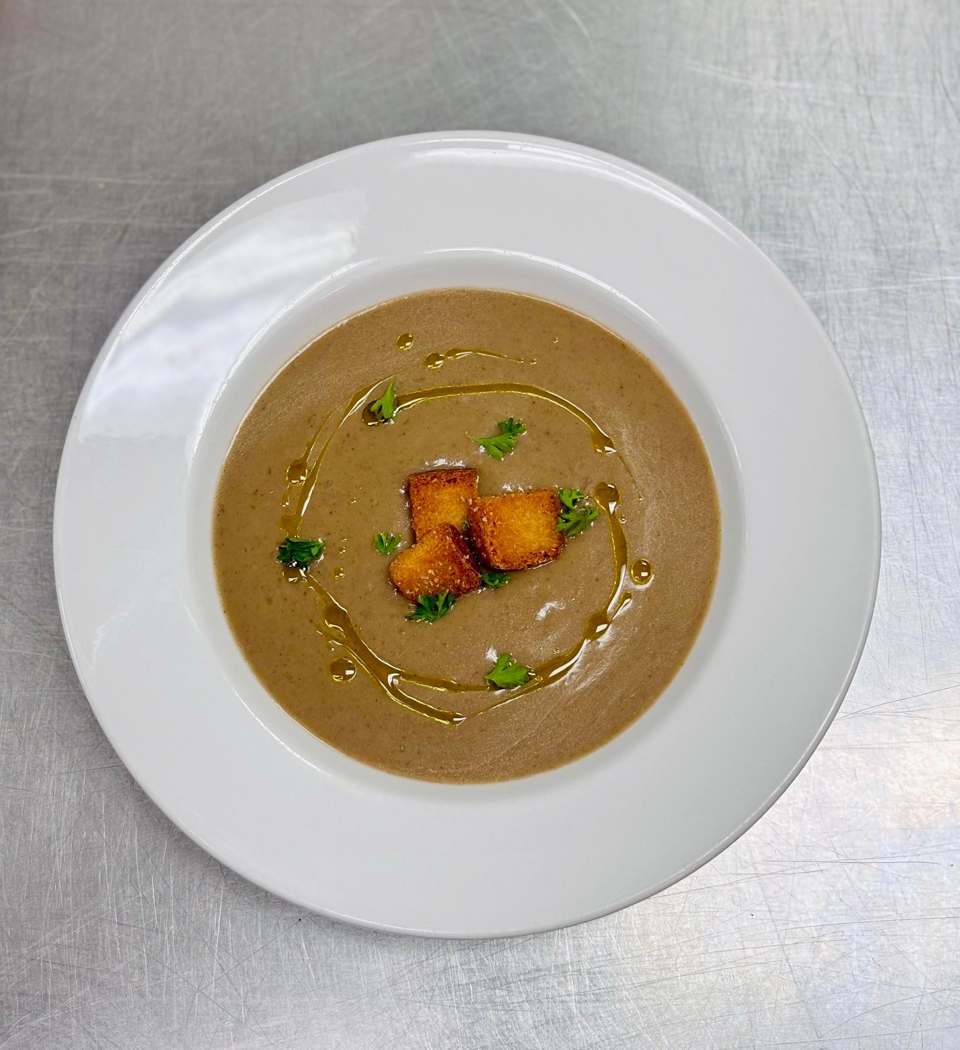 Creamy brown soup in a white bowl, garnished with croutons, parsley, and olive oil drizzle on a metal surface.