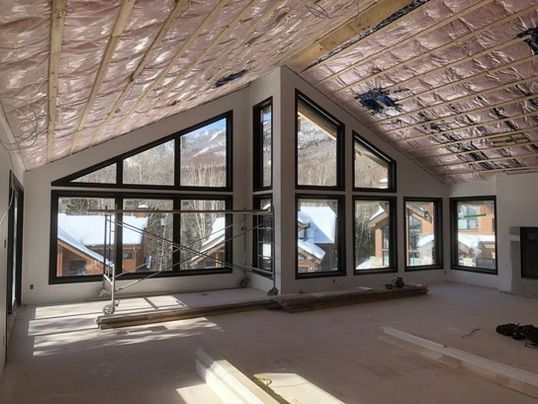 Intérieur d'une maison en construction avec de grandes fenêtres et une isolation. Neige visible à l'extérieur.