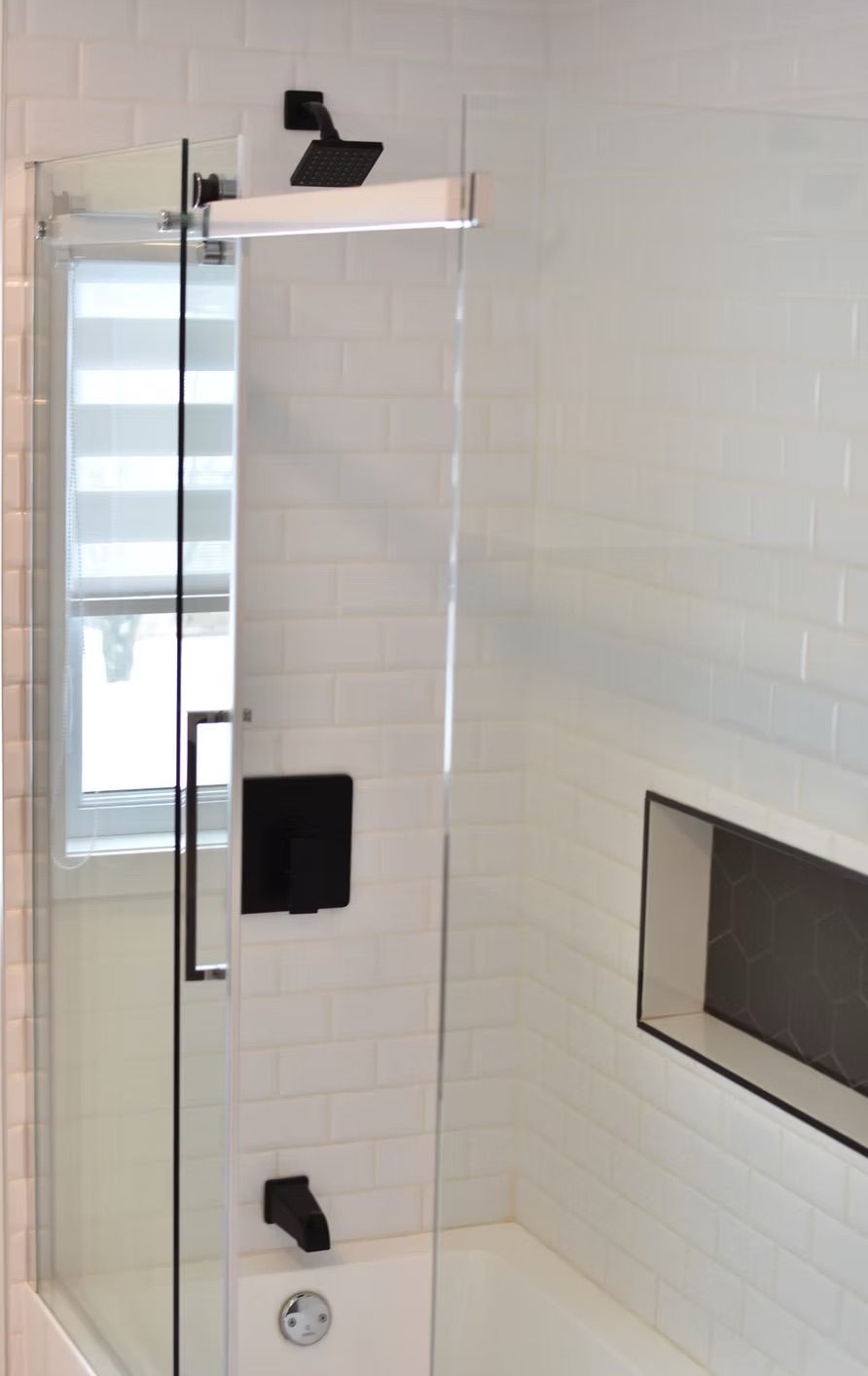 Salle de bains moderne avec porte de douche en verre, robinetterie noire, carrelage métro blanc et étagère encastrée.