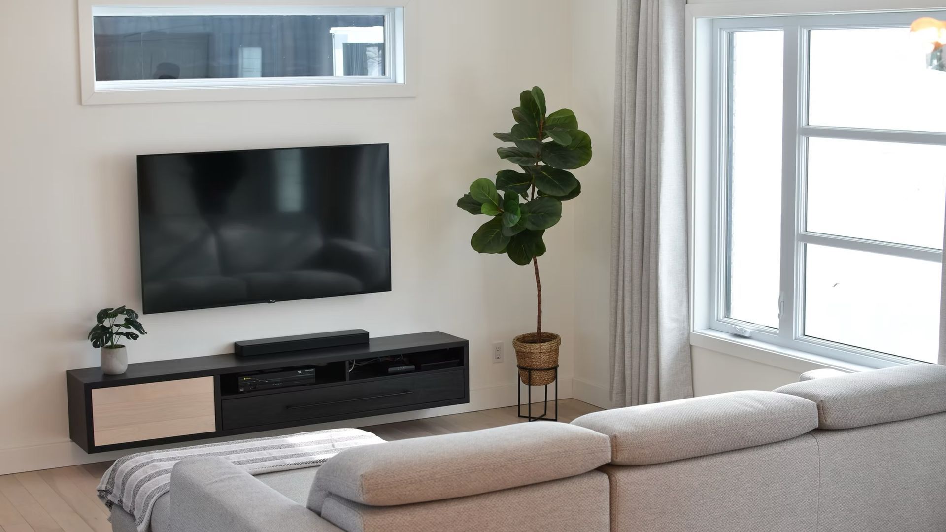 Salon moderne avec télévision suspendue, barre de son et canapé d'angle près d'une grande fenêtre et d'un figuier lyre.