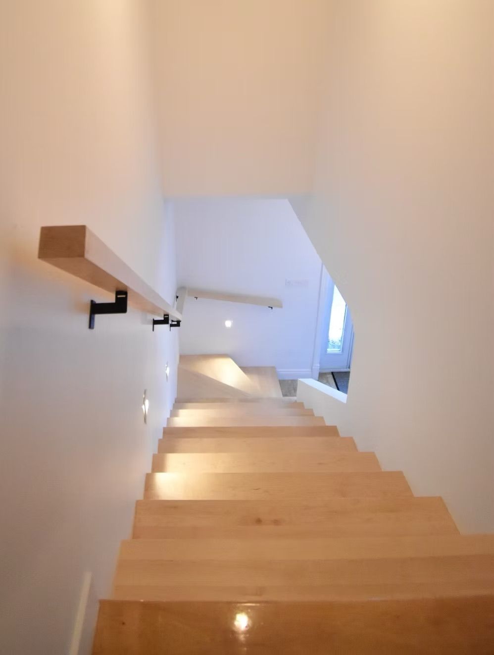 Escalier en bois avec rampe à gauche, menant vers l'étage, avec éclairage encastré au mur.