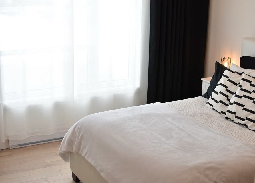 Chambre avec lit blanc, coussins à motifs, voilages et rideaux noirs.