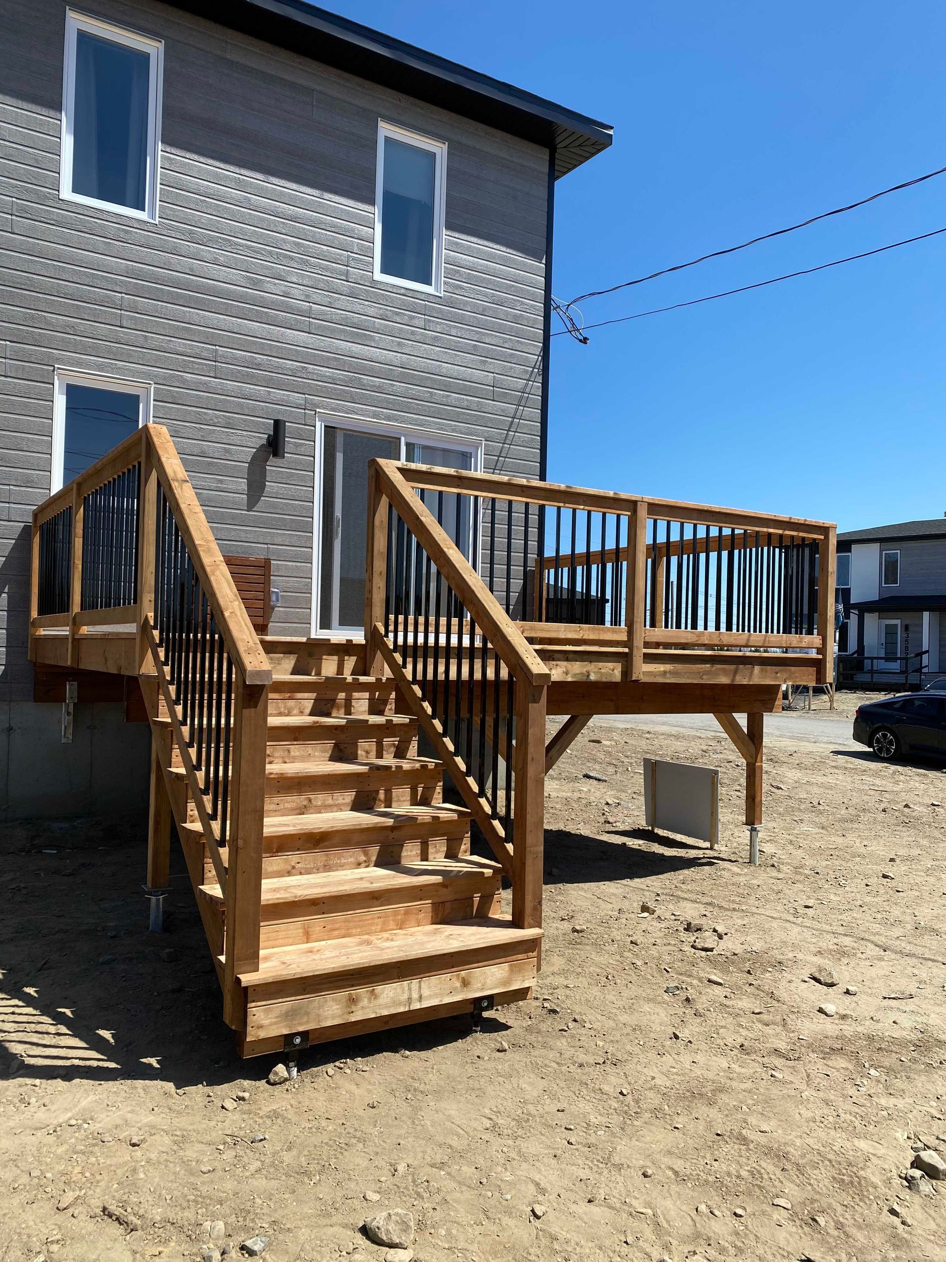 Terrasse en bois avec escalier attenant à une maison grise, rambarde marron et balustres noirs, par une journée ensoleillée.