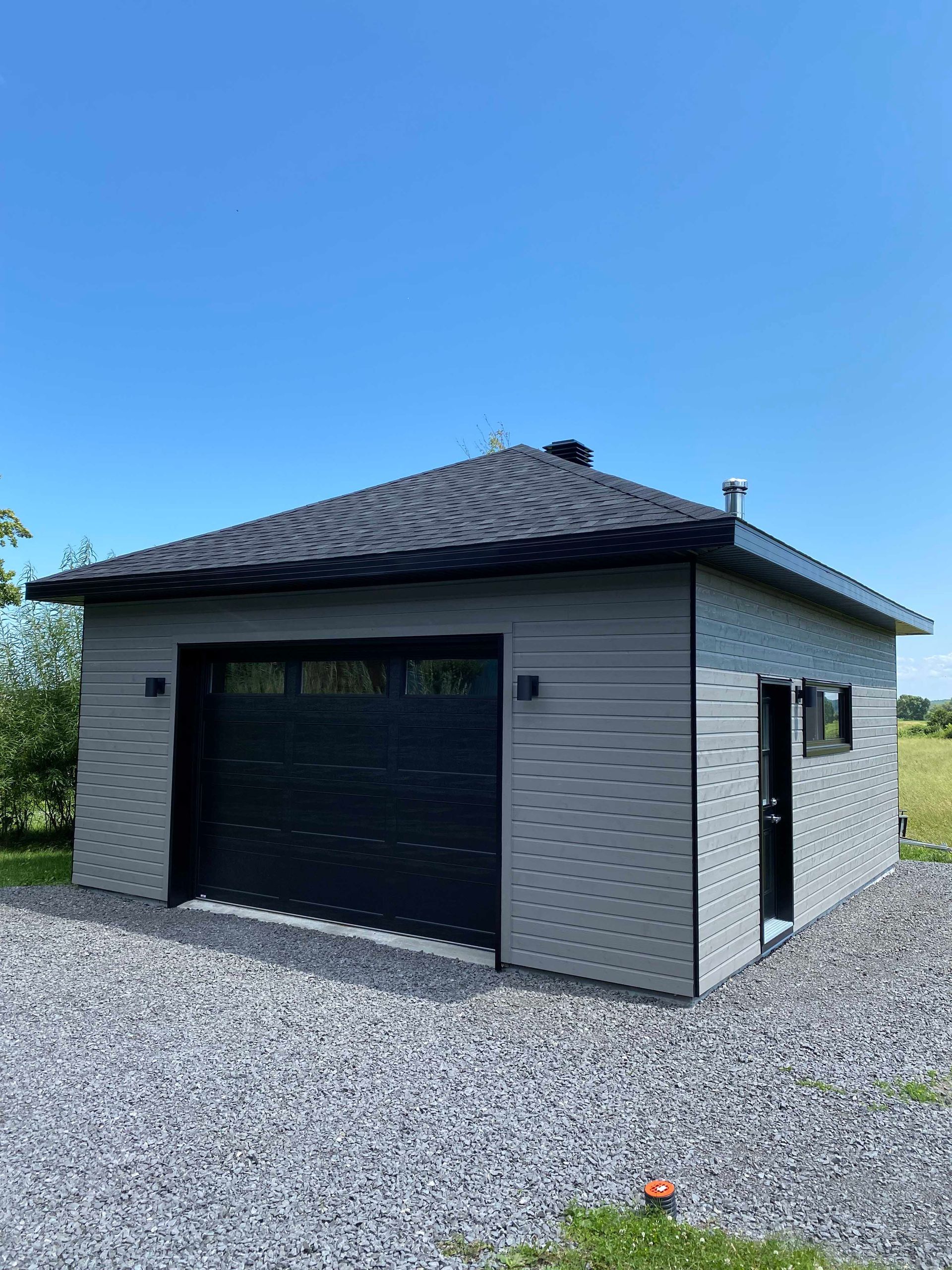 Garage gris avec porte et toit noirs, sur une allée de gravier sous un ciel bleu.