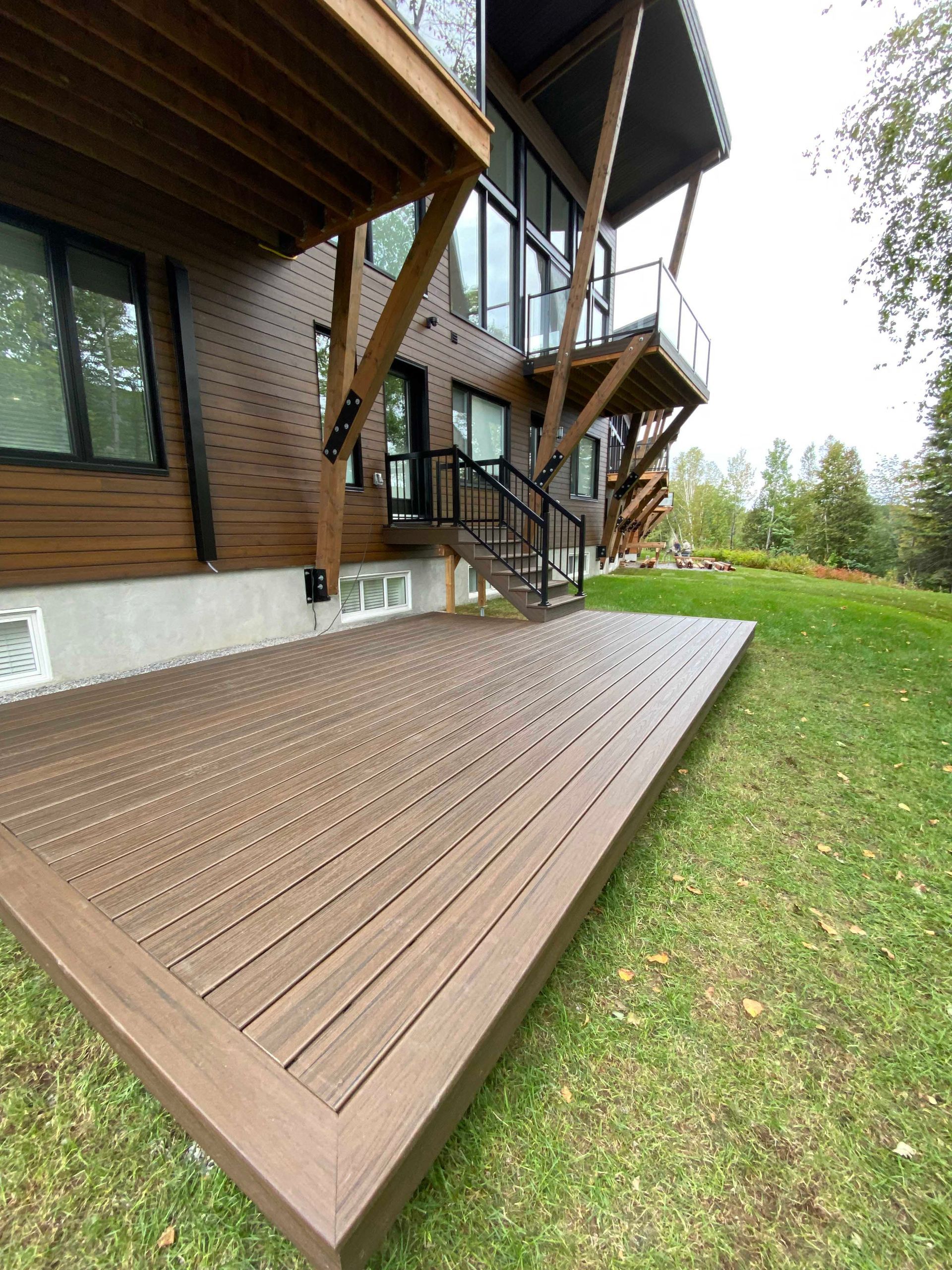Terrasse en composite marron devant une maison moderne avec terrasse surélevée, escalier et pelouse verte.
