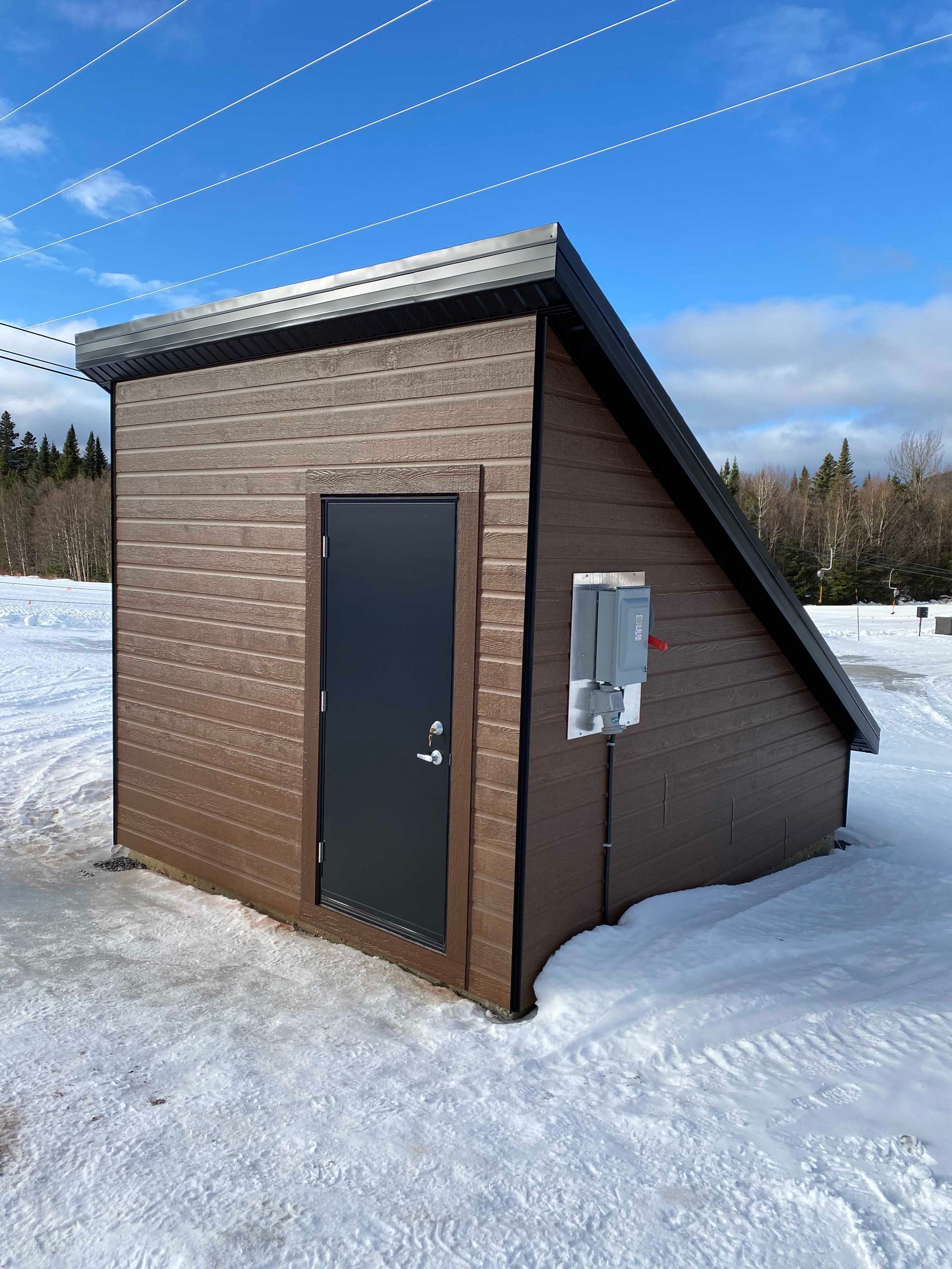 Abri utilitaire marron dans un paysage enneigé, avec une porte et un tableau électrique noirs.