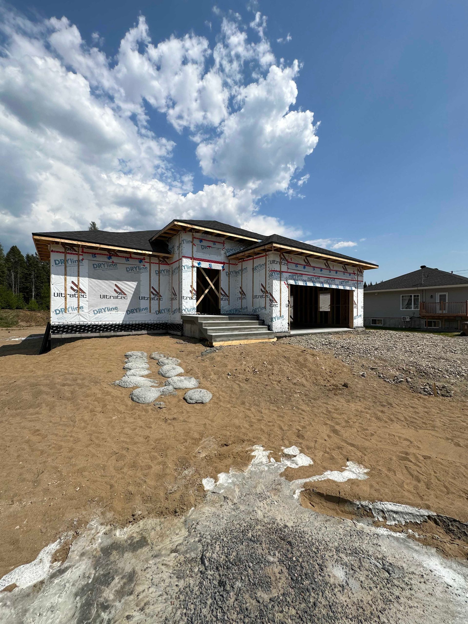 Maison en construction par une journée ensoleillée ; murs extérieurs recouverts d'un film blanc, ciel bleu nuageux.