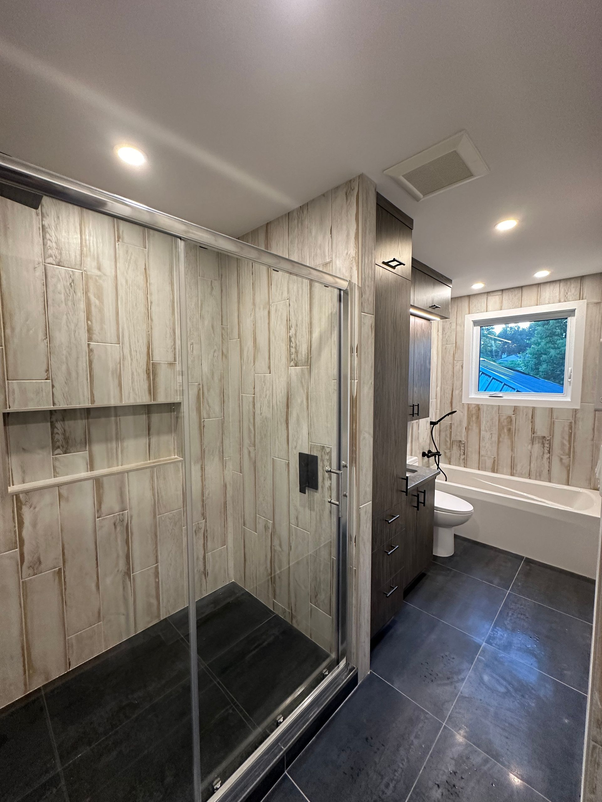 Salle de bains avec panneaux muraux imitation bois, douche en verre, carrelage gris au sol et baignoire blanche.