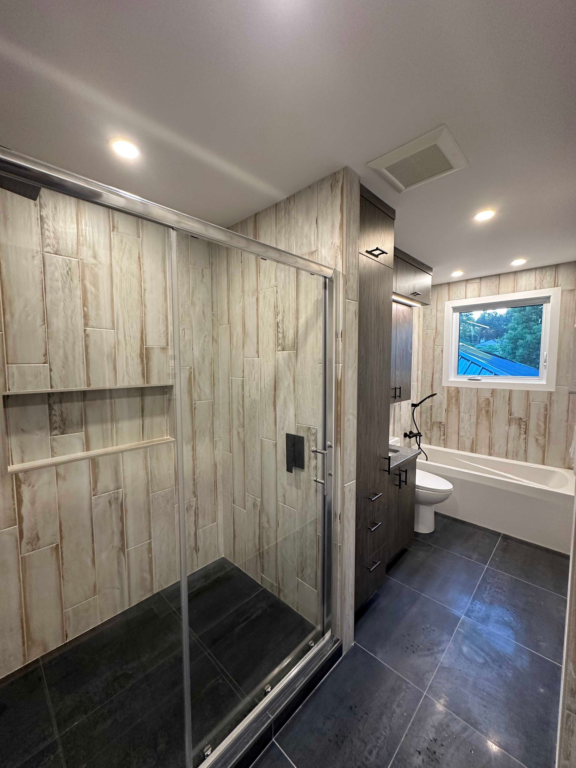 Salle de bains avec murs carrelés imitation bois, douche en verre, sol carrelé foncé, meuble-lavabo, toilettes et baignoire.