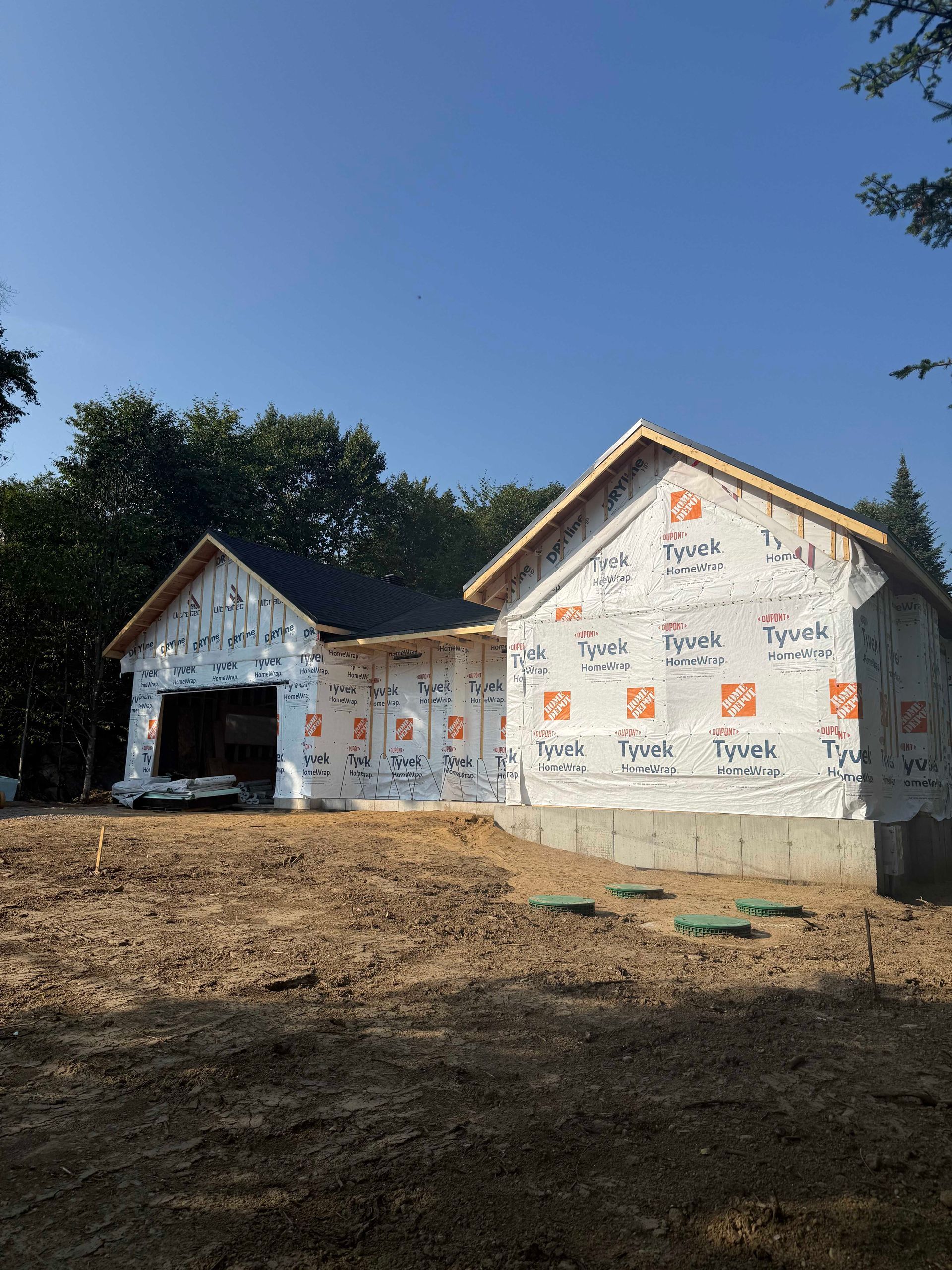 Maison en construction ; murs extérieurs recouverts de Tyvek, ossature bois. Ciel bleu, arbres verts en arrière-plan.