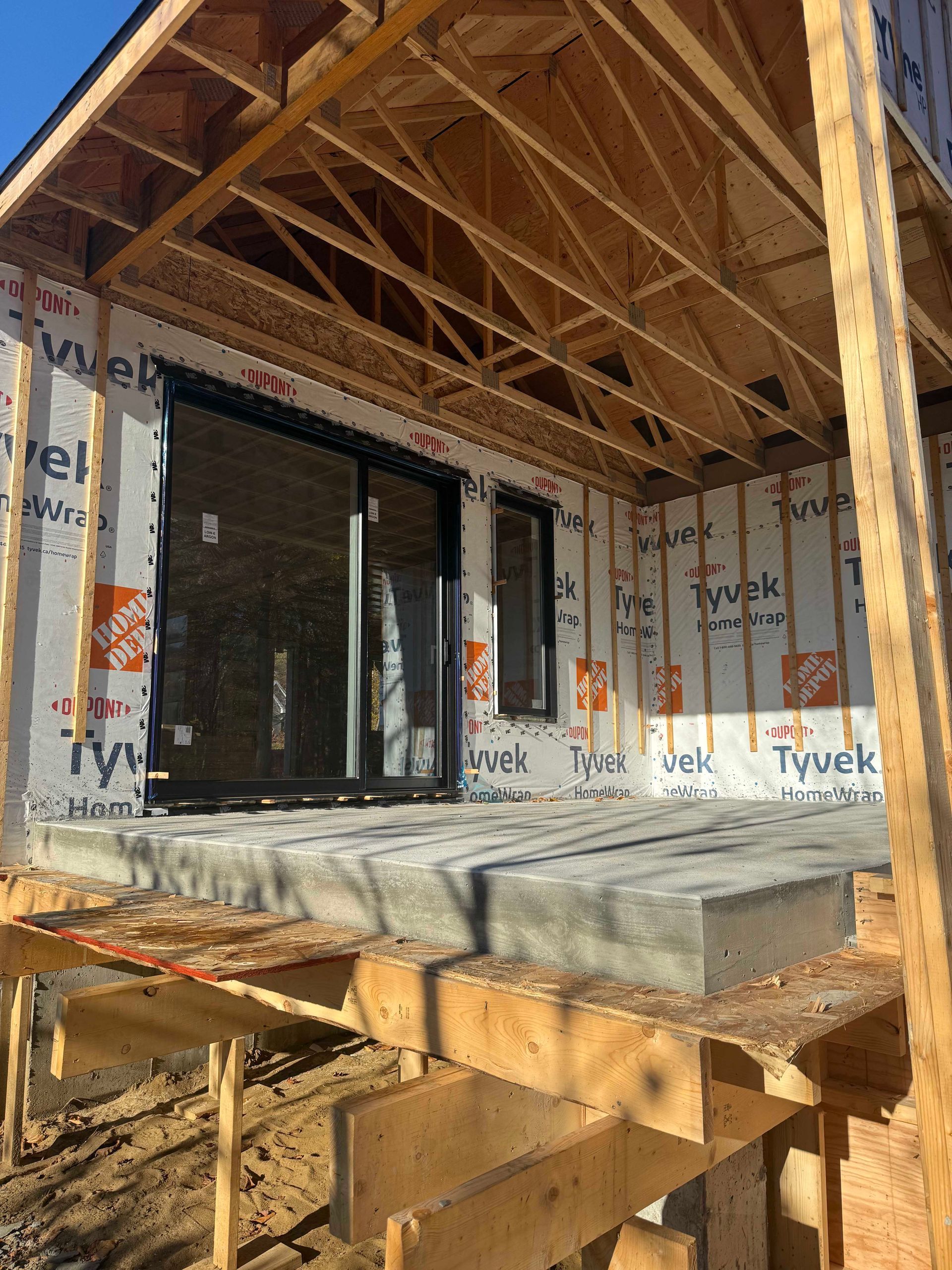 Chantier de construction d'une maison avec charpente apparente en bois, terrasse en béton et portes coulissantes en verre à cadre noir.