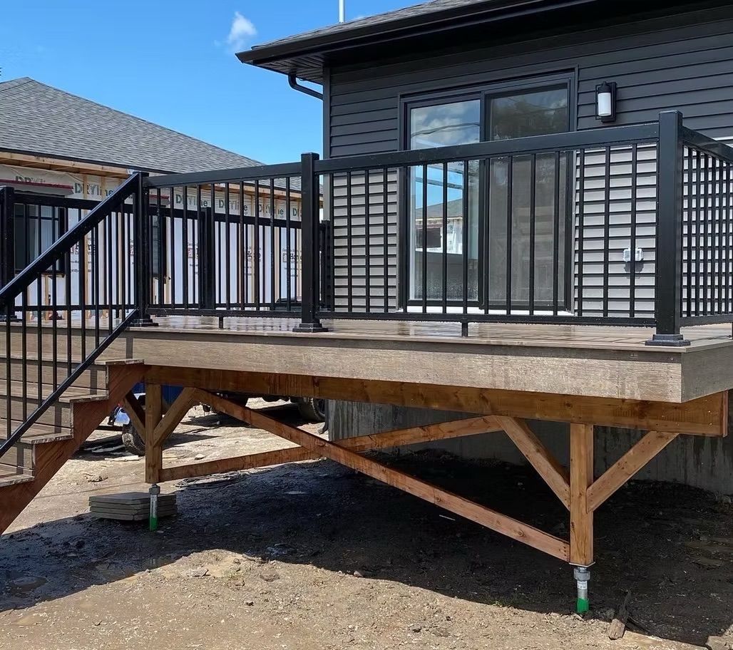 Une terrasse à balustrade noire avec escalier, soutenue par des poutres en bois, se détache sur la façade grise d'une maison, par une journée ensoleillée.