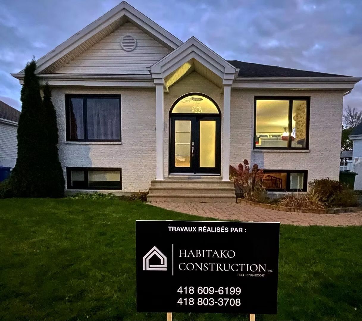 Une maison en briques blanches avec des boiseries noires, une porte éclairée et un panneau « HALITAKO CONSTRUCTION » sur la pelouse.