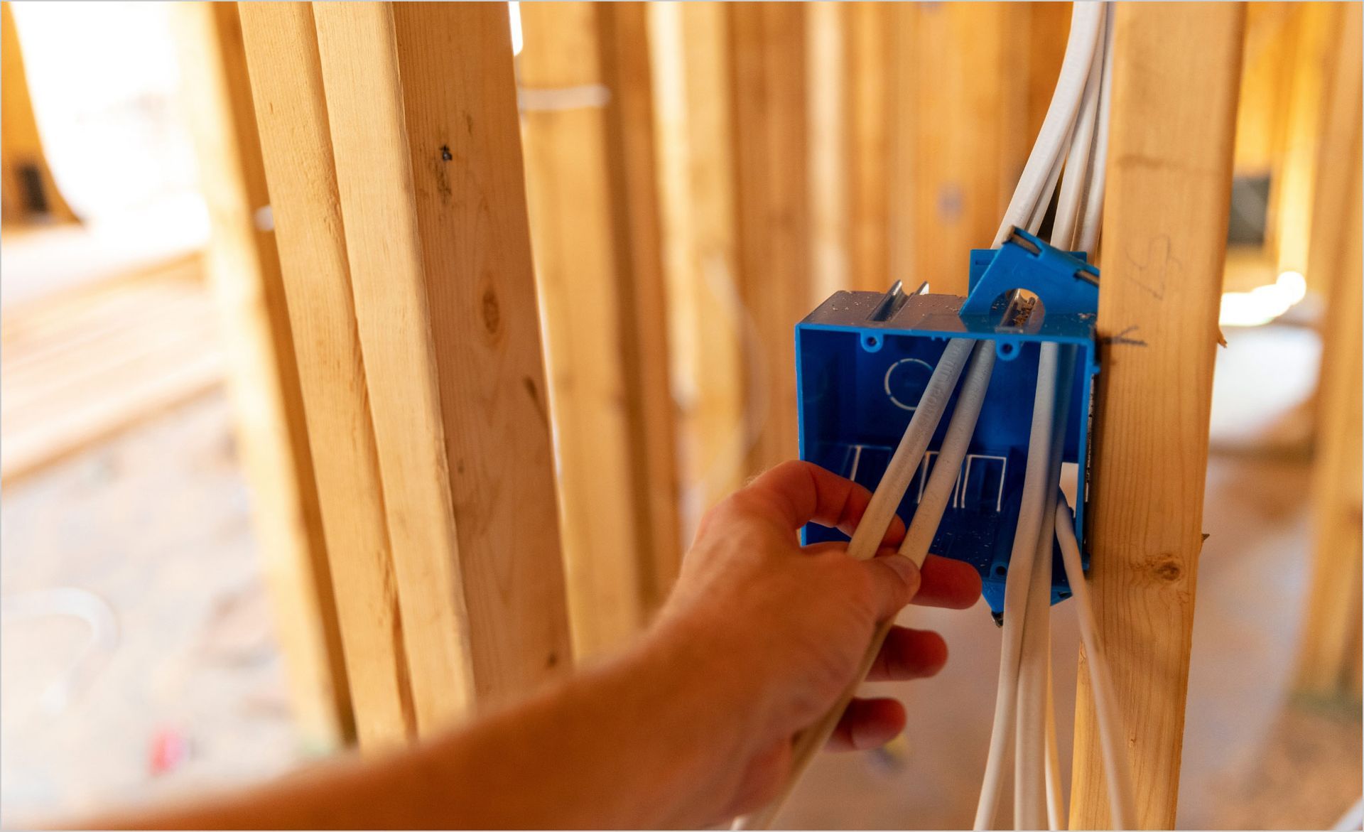 Hand fastening blue electrical box into wooden wall framing with wires nearby
