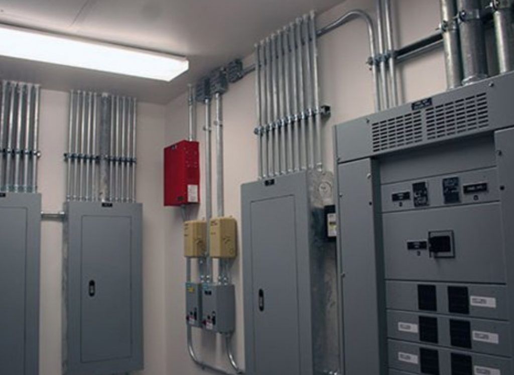 Electrical panel room with gray cabinets, conduit pipes, and a red fire box on the wall