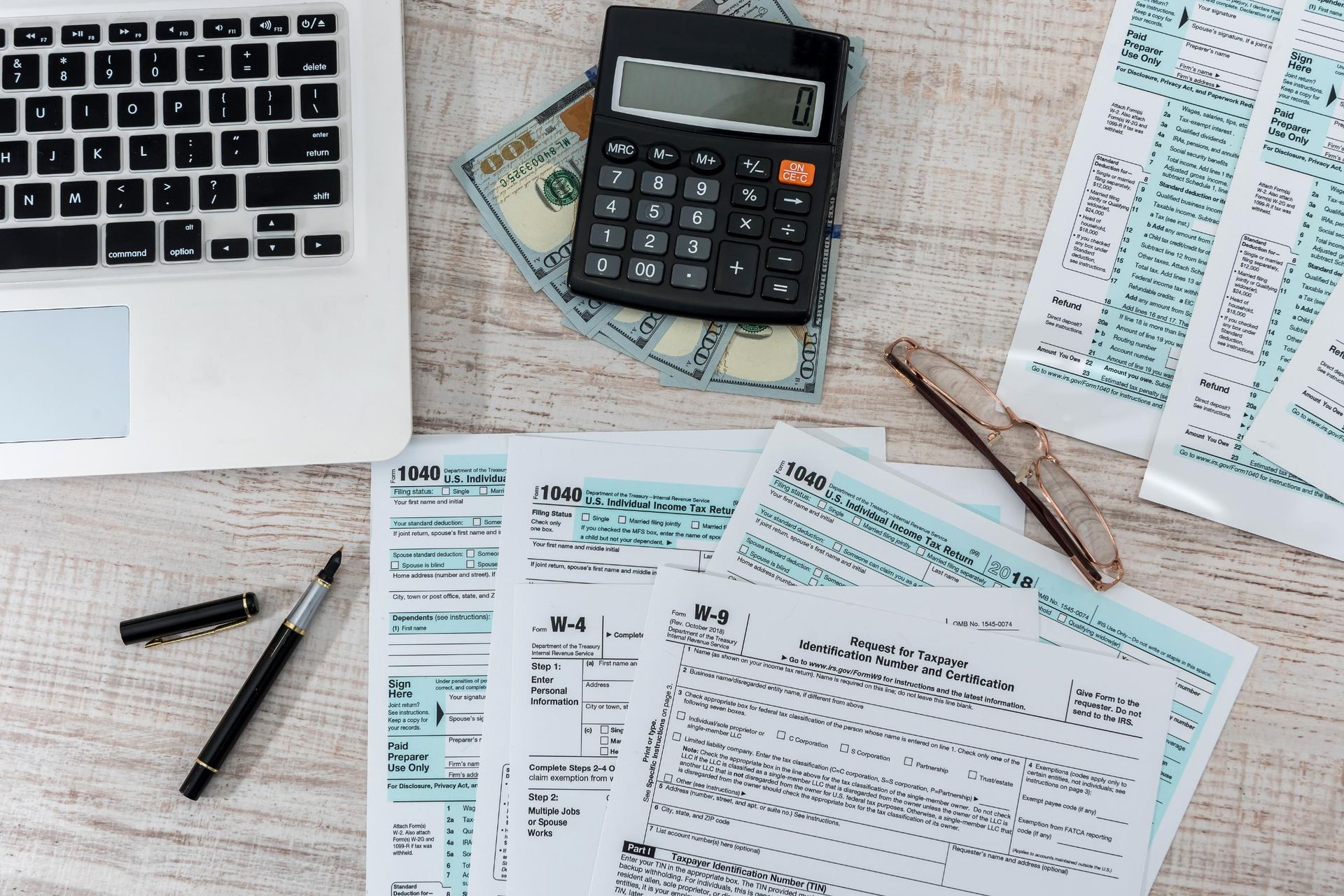 Laptop, tax forms, calculator, cash, and pen on a wooden desk, suggesting financial paperwork.