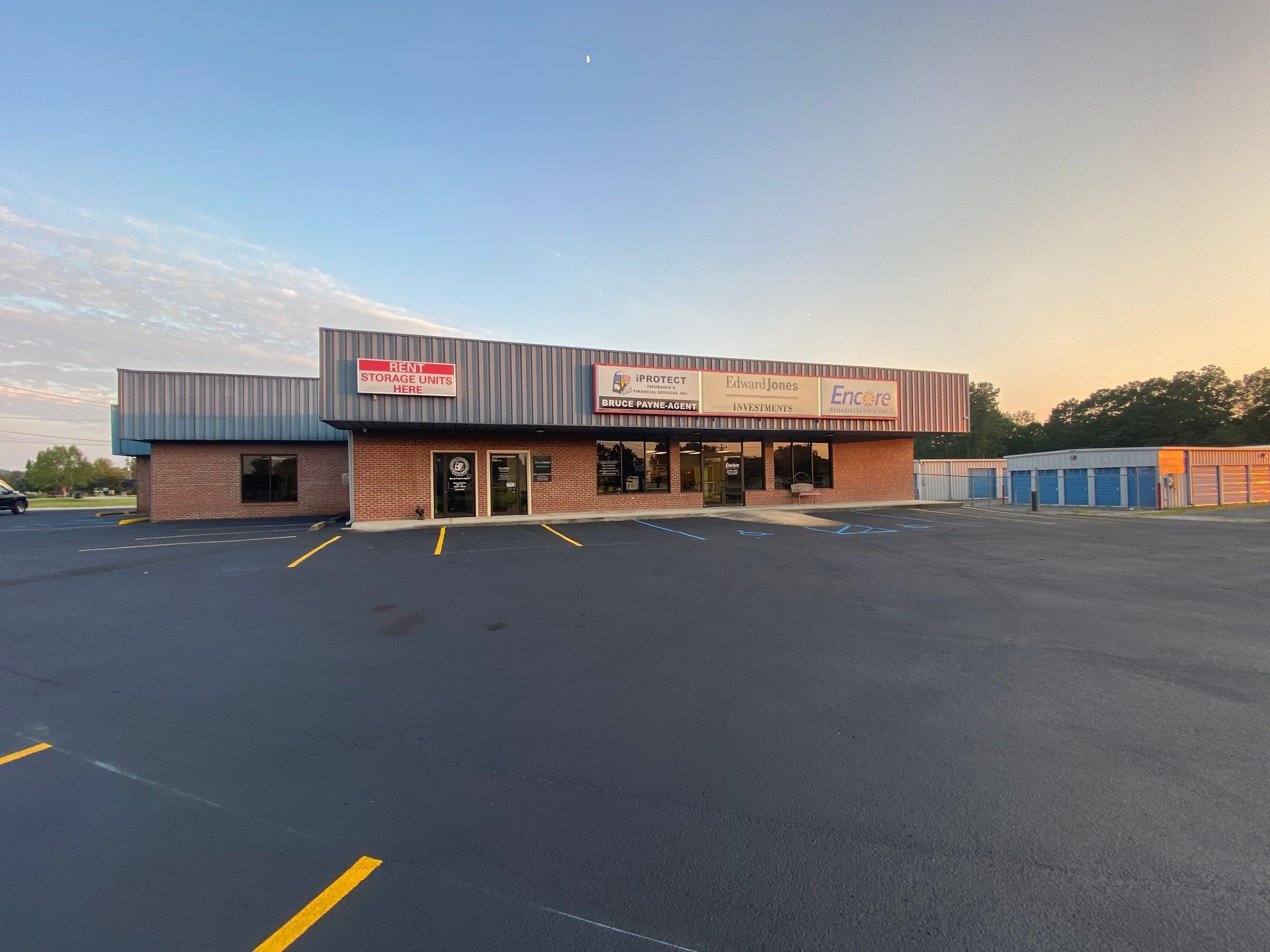 Storefront with signs, brick facade, and a large paved parking lot under a blue sky.