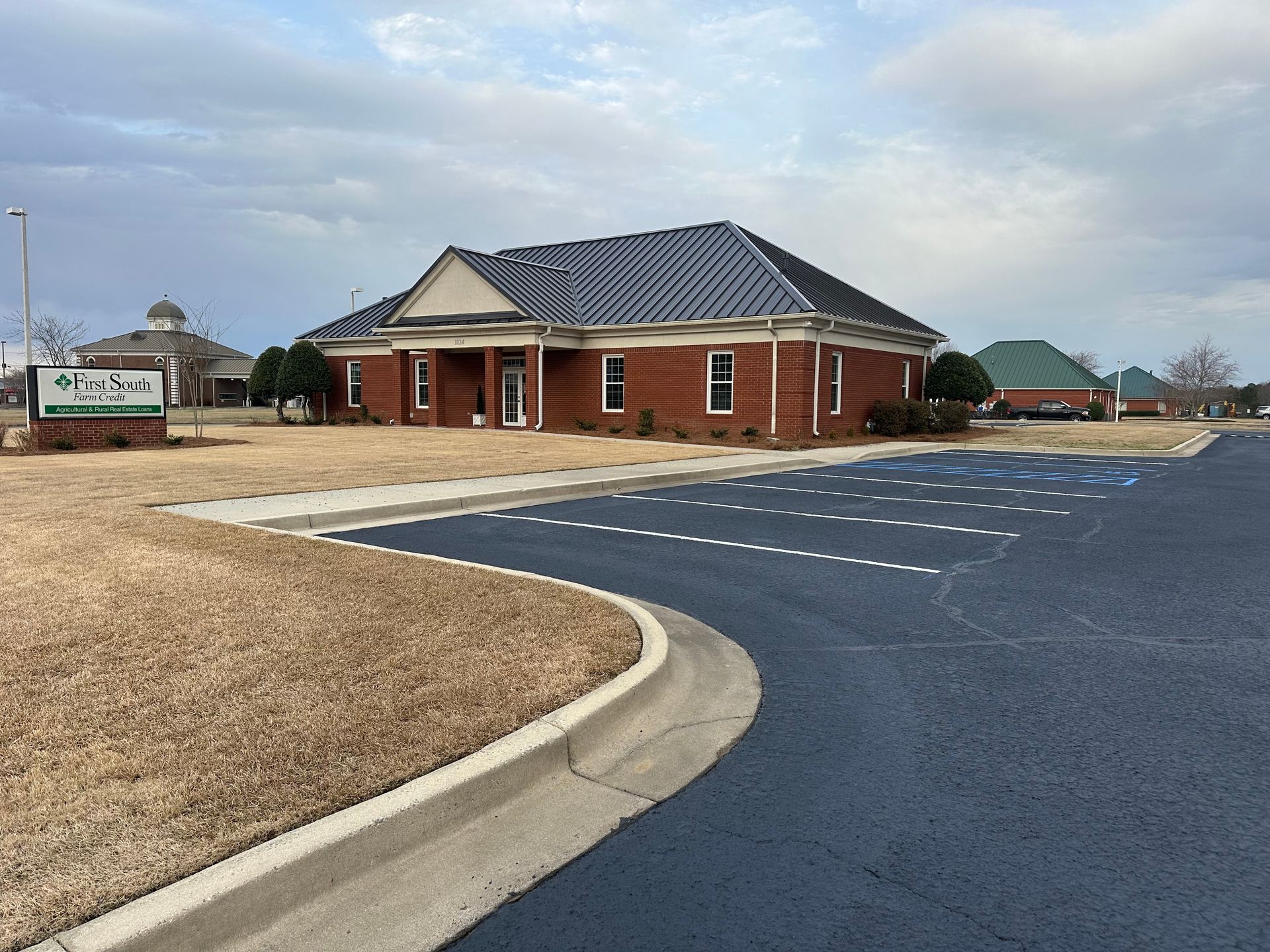 A brick building with a dark roof and a paved parking lot on a sunny day.
