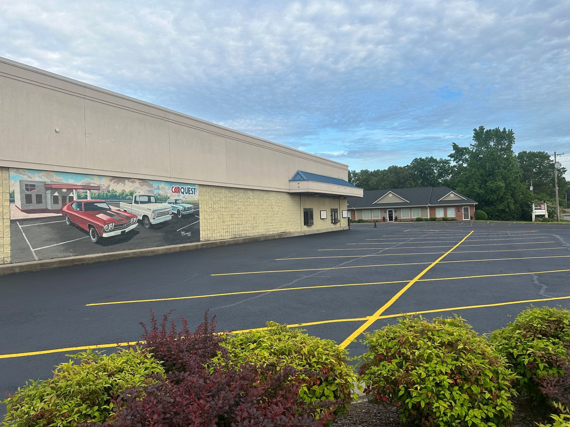 Exterior of a building with a mural of classic cars on the side, empty parking lot, and bushes in the foreground.
