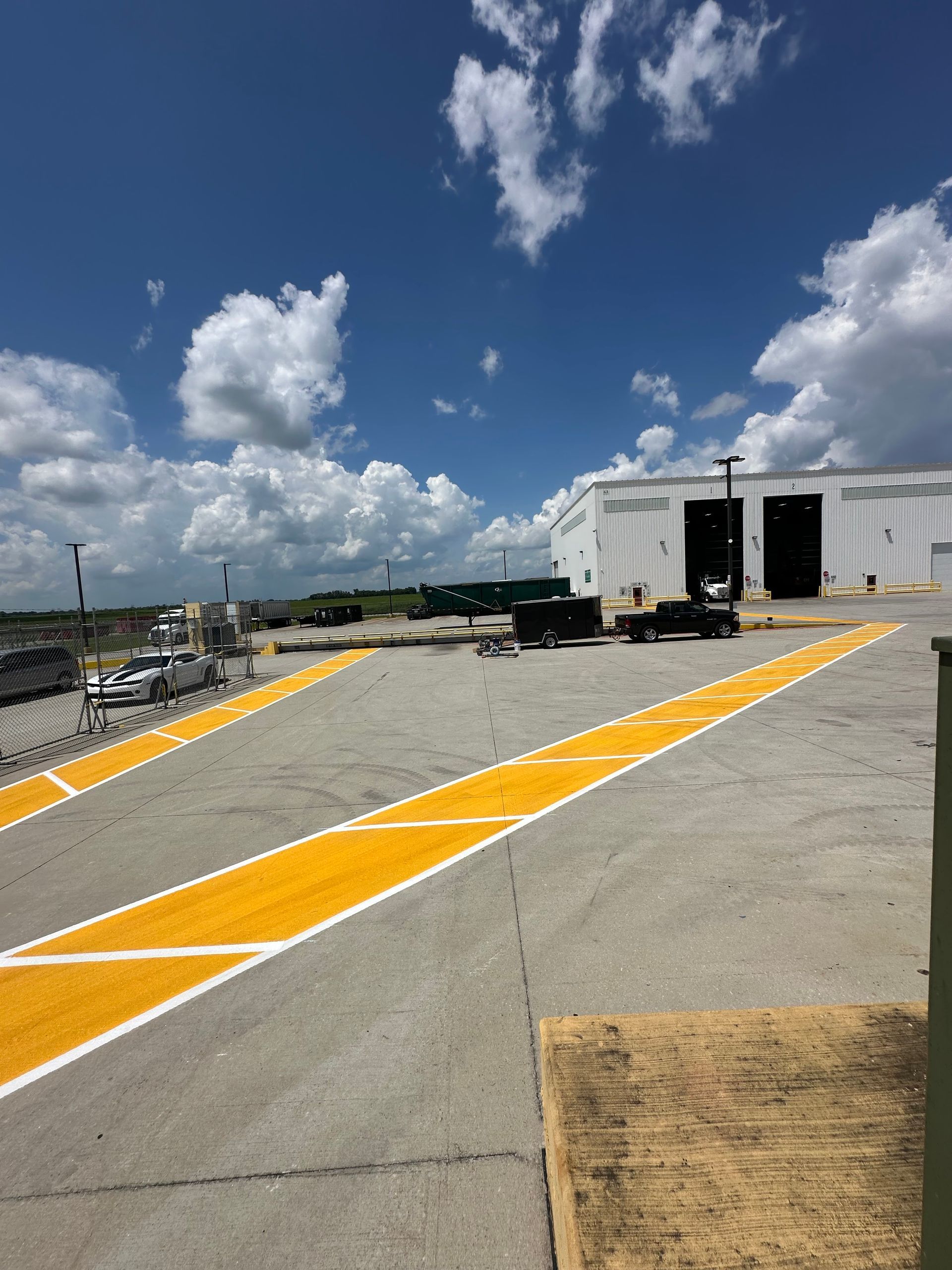 Yellow painted lines on gray concrete. Large white building with open bay doors. Blue sky, clouds.