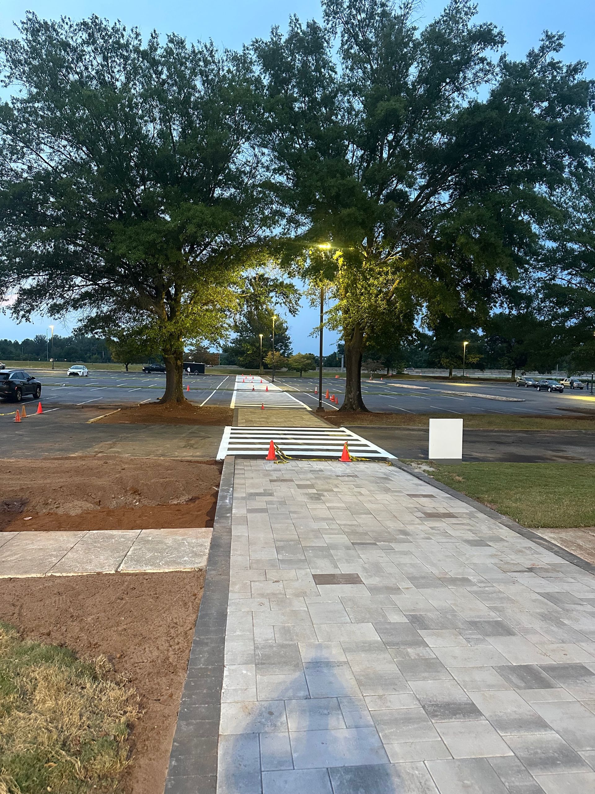 Stone walkway leading between trees towards a parking area. Construction barriers and lights are visible.
