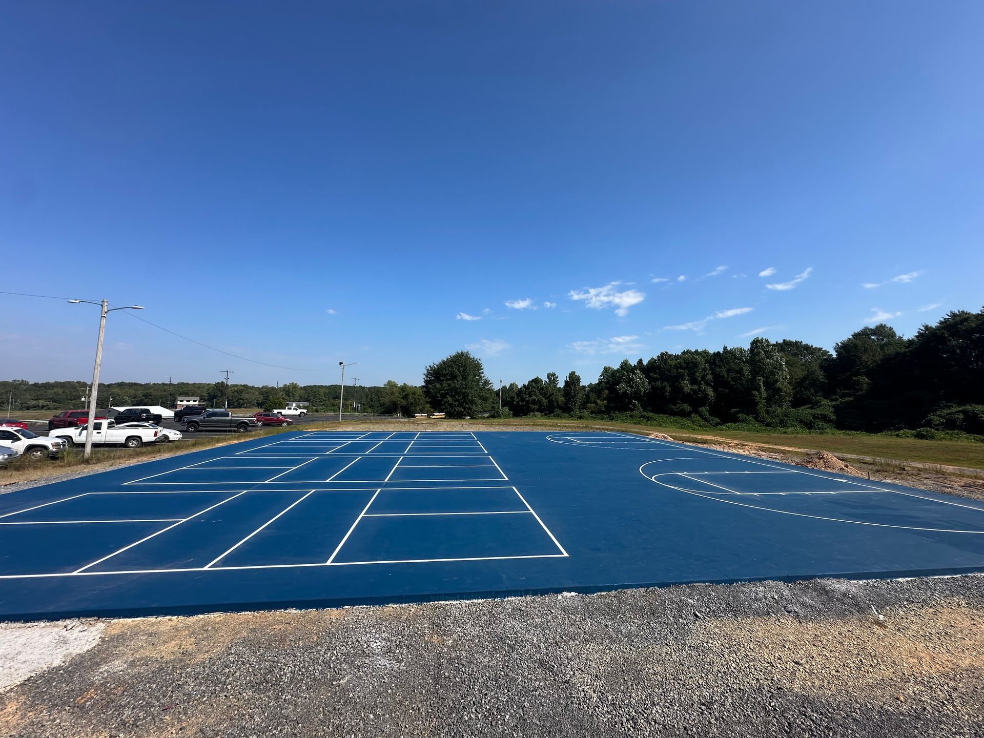 Blue outdoor sports court with white lines, parked cars, and trees under a blue sky.