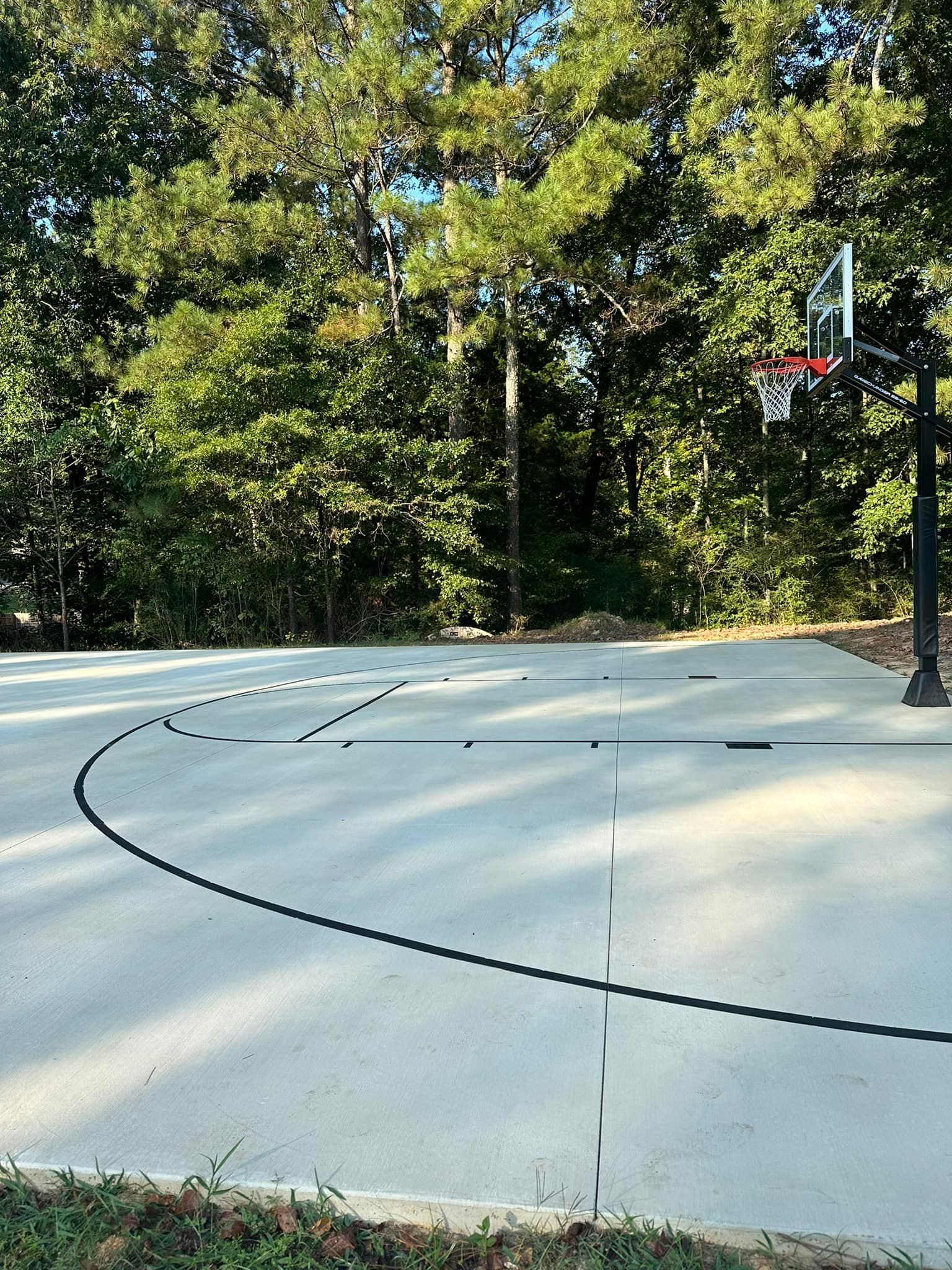 Outdoor basketball court with black lines, hoop, and trees in background.