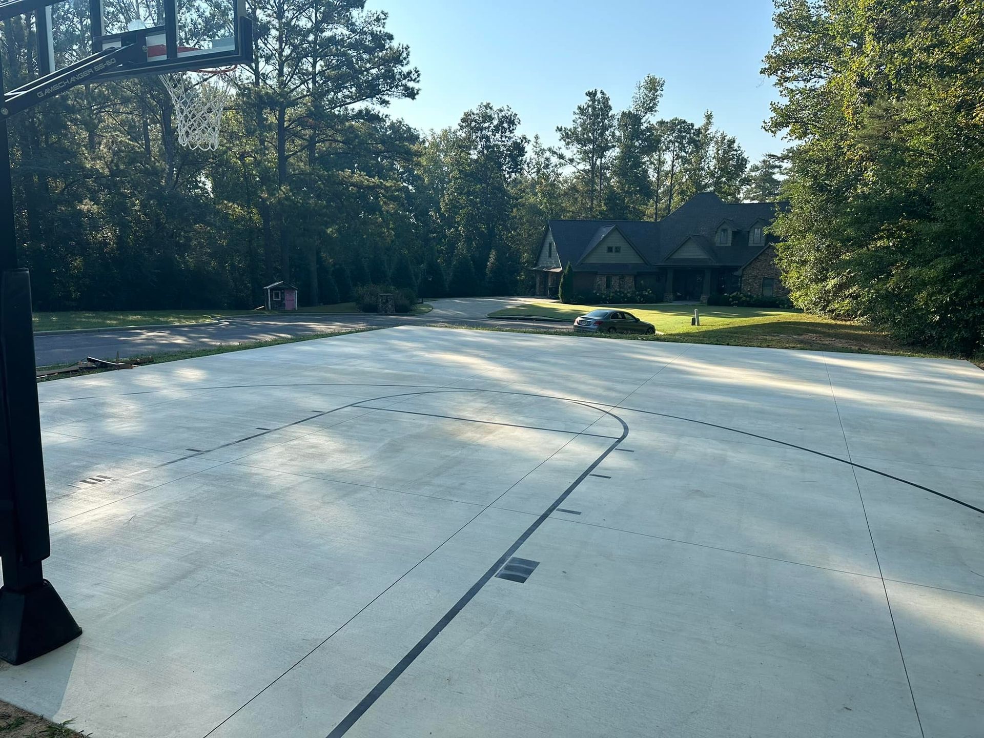 Basketball court with hoop in front of a house, surrounded by trees and a clear sky.