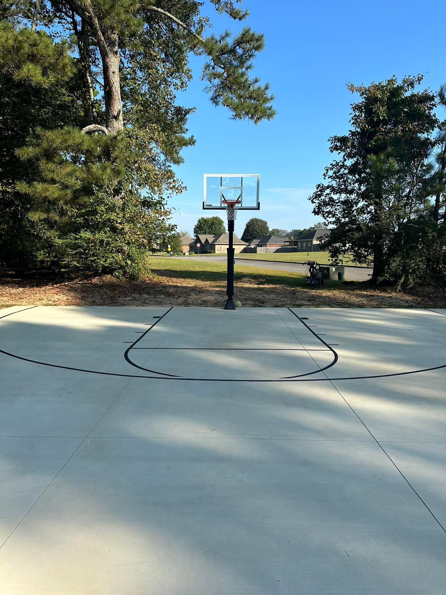 Basketball hoop on a concrete court, trees frame the view, a clear sky overhead.