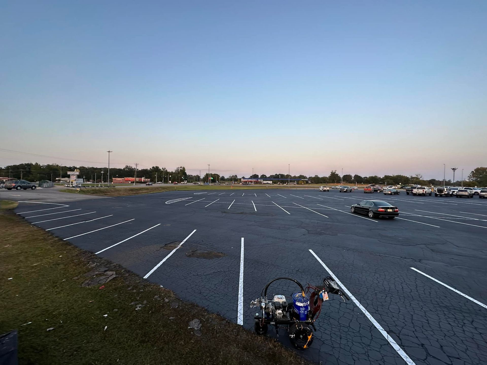 Parking lot with freshly painted white lines; painting machine in foreground; cars parked in the distance; dusk sky.