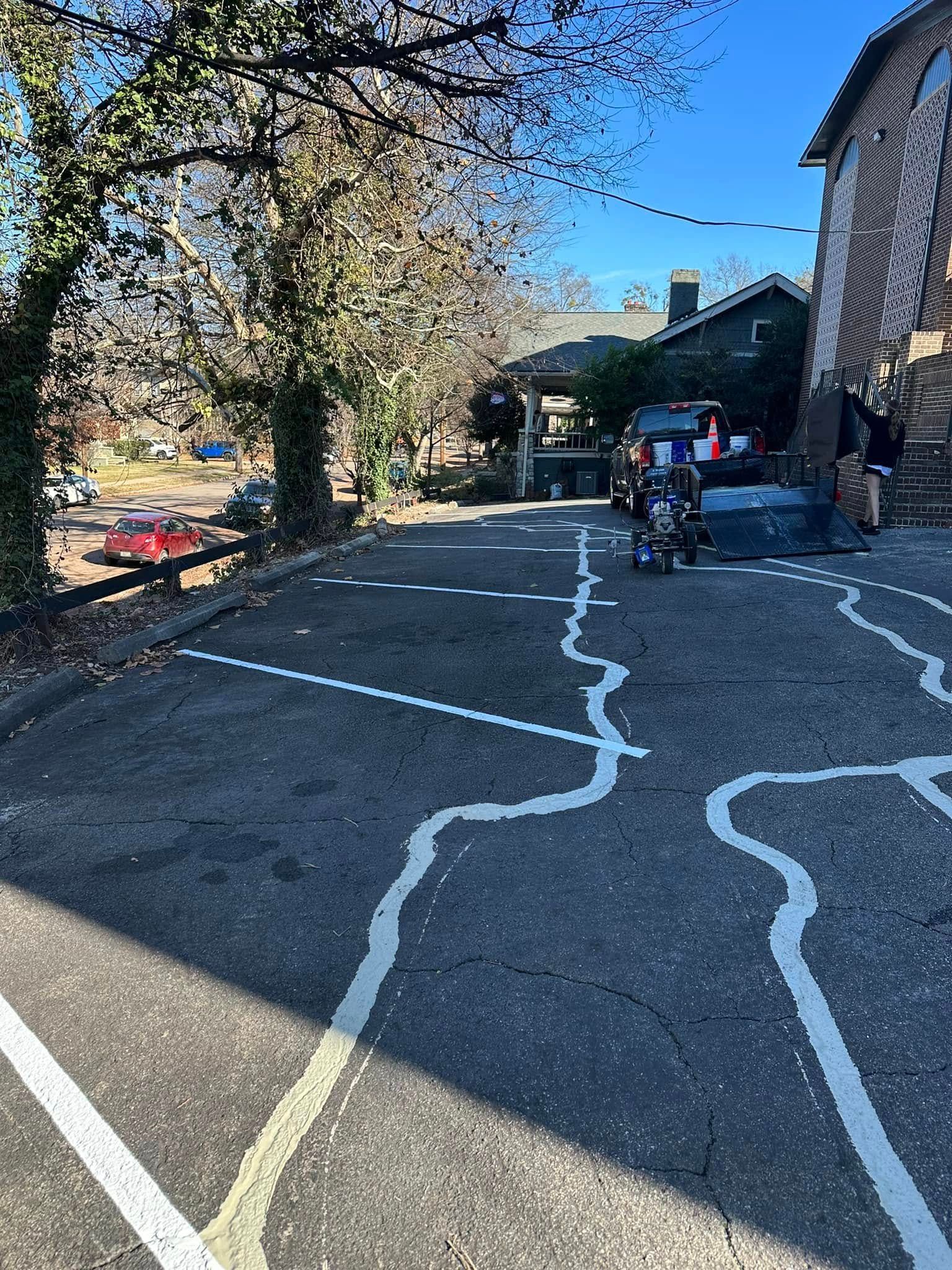 Asphalt parking lot with white painted lines. Trees and a brick building on right. Cars parked.