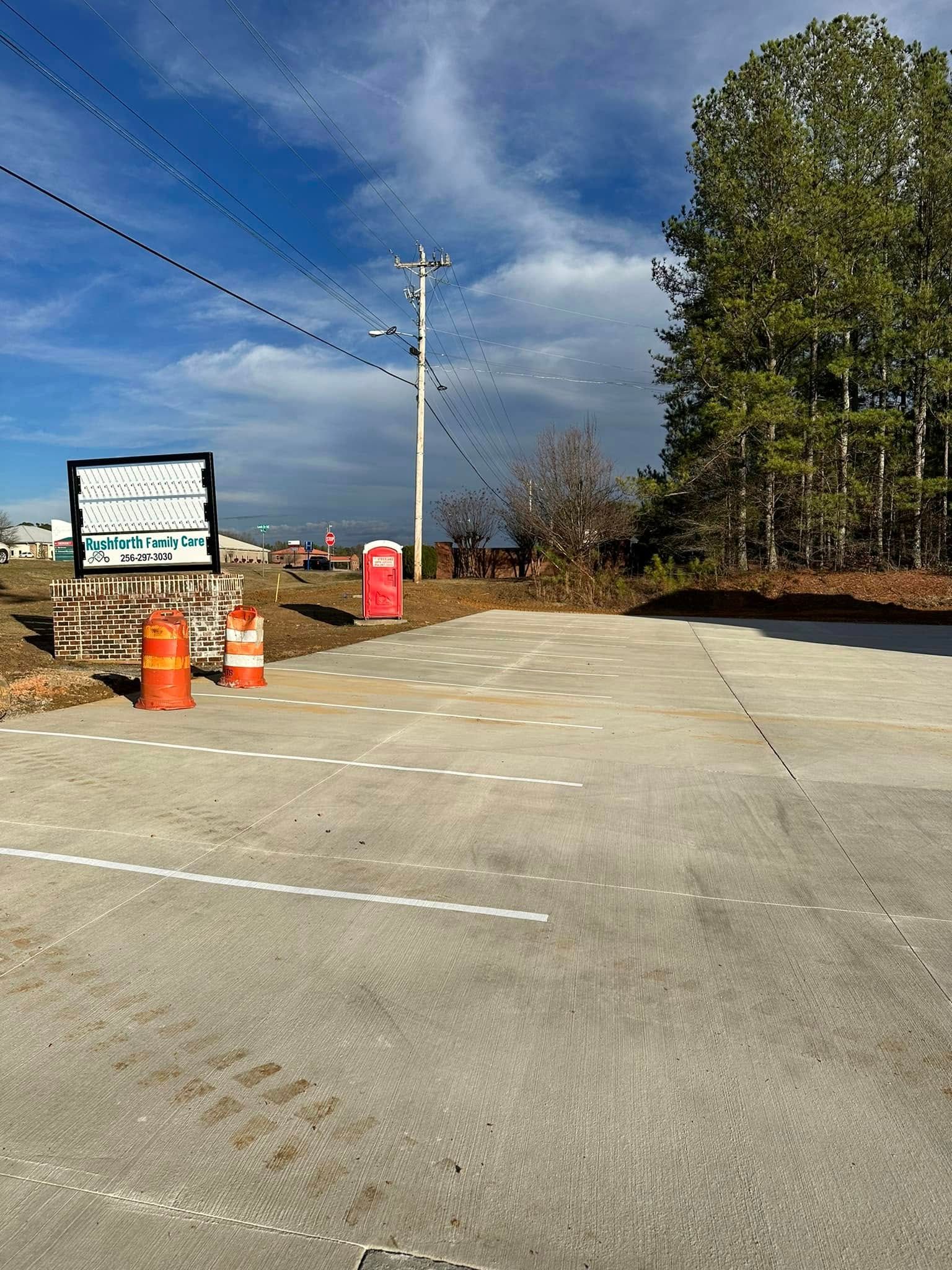 Concrete parking area with power lines, porta-potty, construction cones, and sign. Trees and cloudy sky in background.