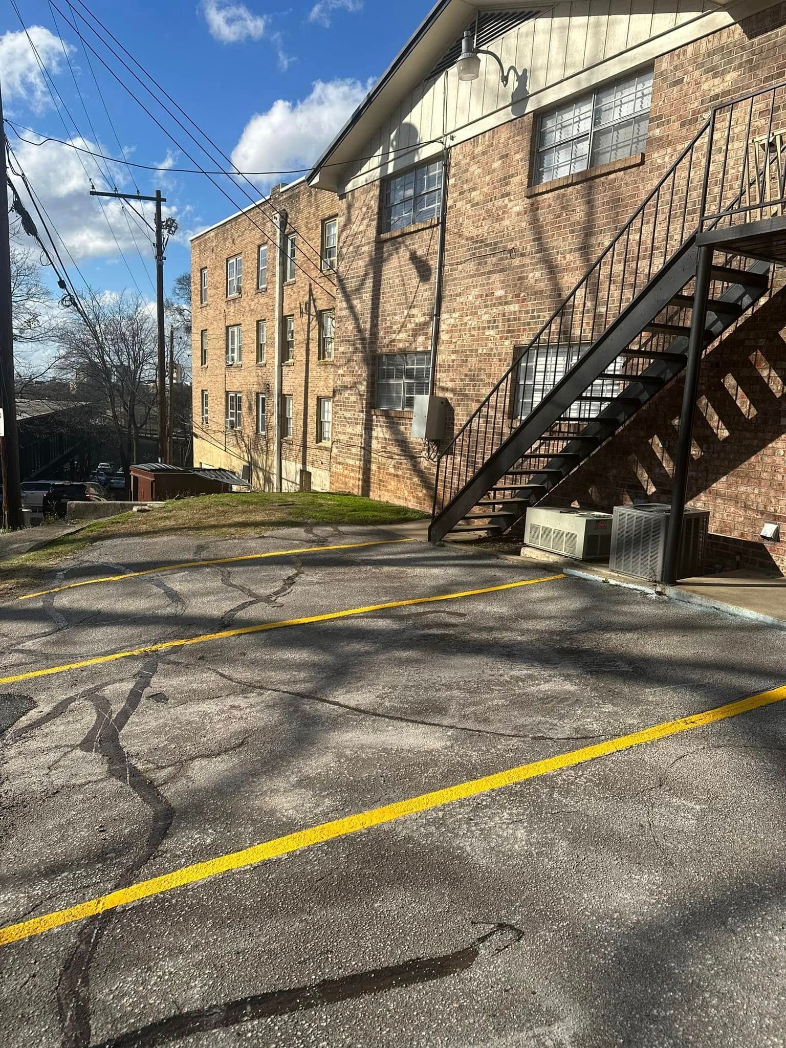 Brick apartment building with exterior metal staircase, adjacent to a cracked asphalt parking lot with yellow lines.