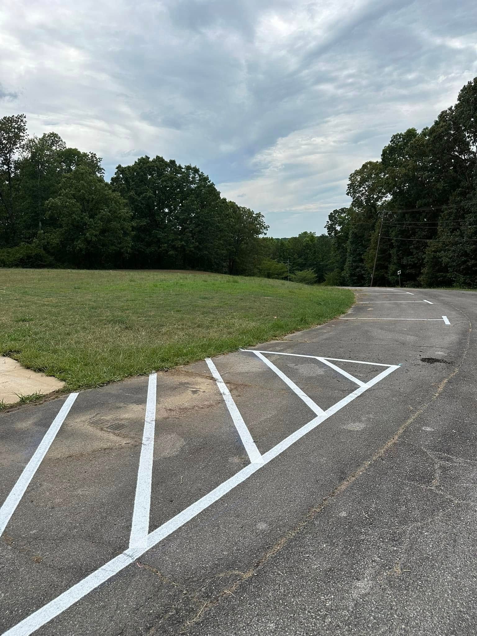 Asphalt driveway with angled parking lines, leading to a grassy area and trees under a cloudy sky.