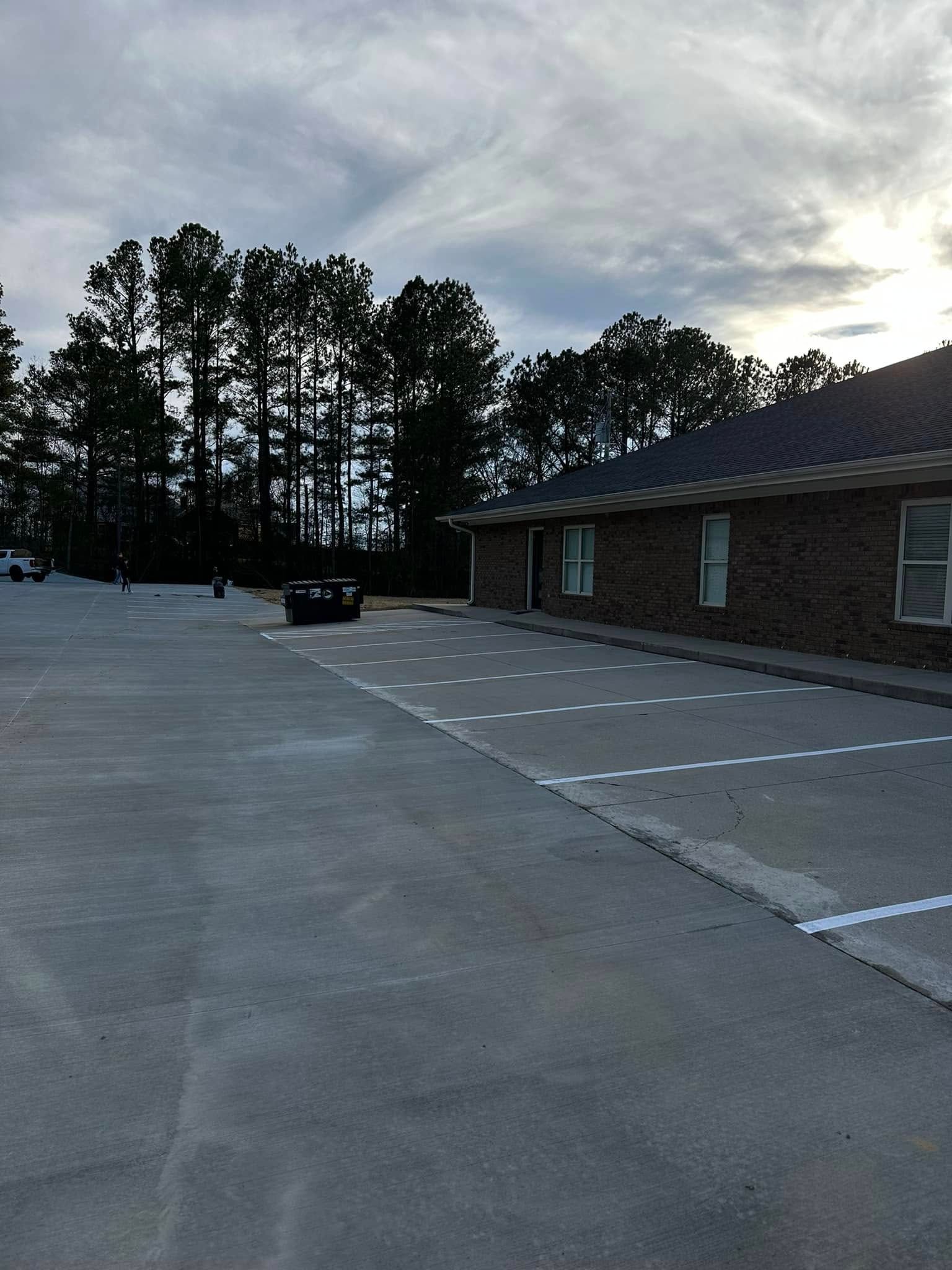 Parking lot with white-lined spaces beside a brick building and trees under a cloudy sky.