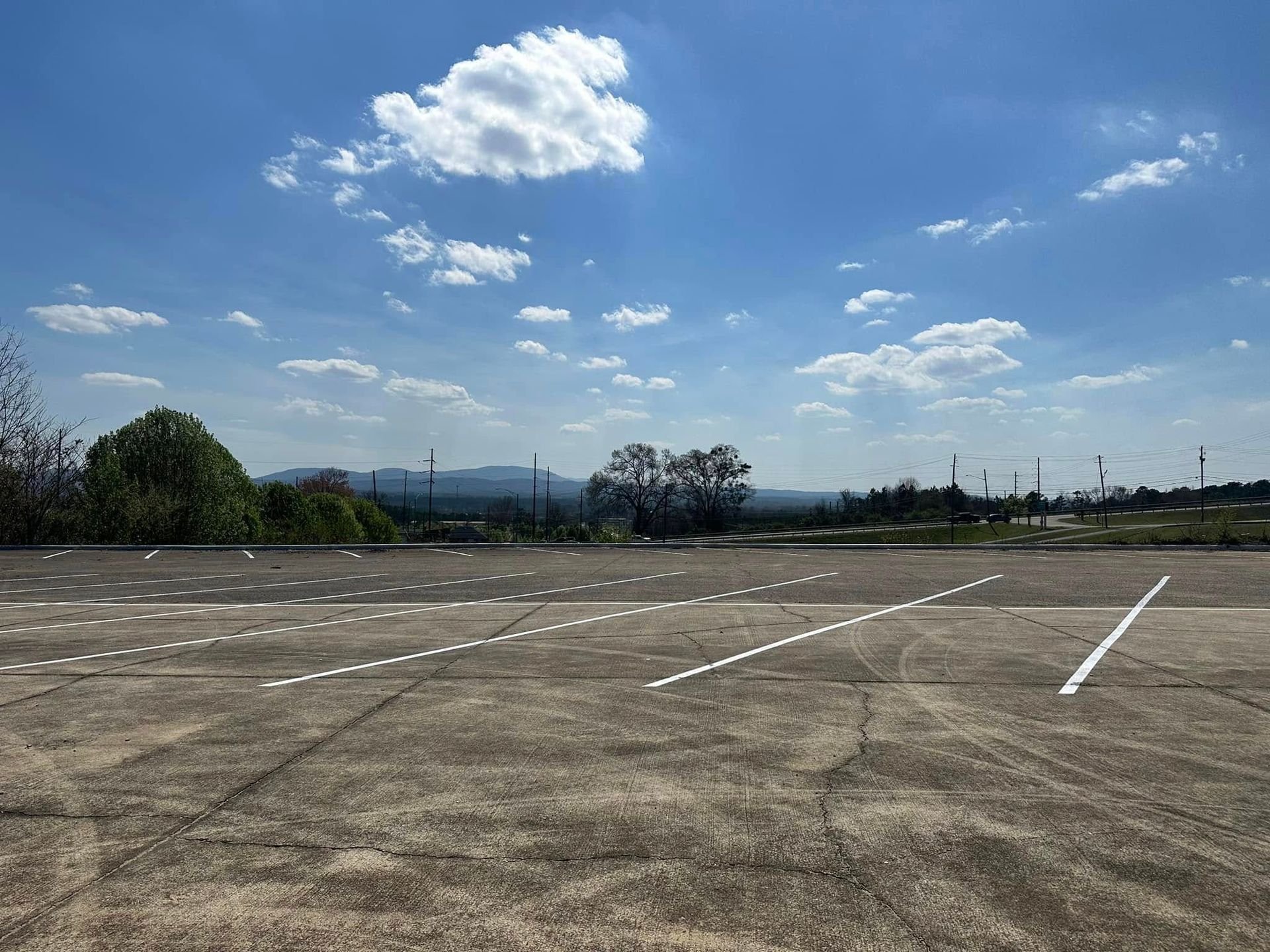 An empty parking lot with faded white lines, trees, and a distant mountain range under a partly cloudy sky.