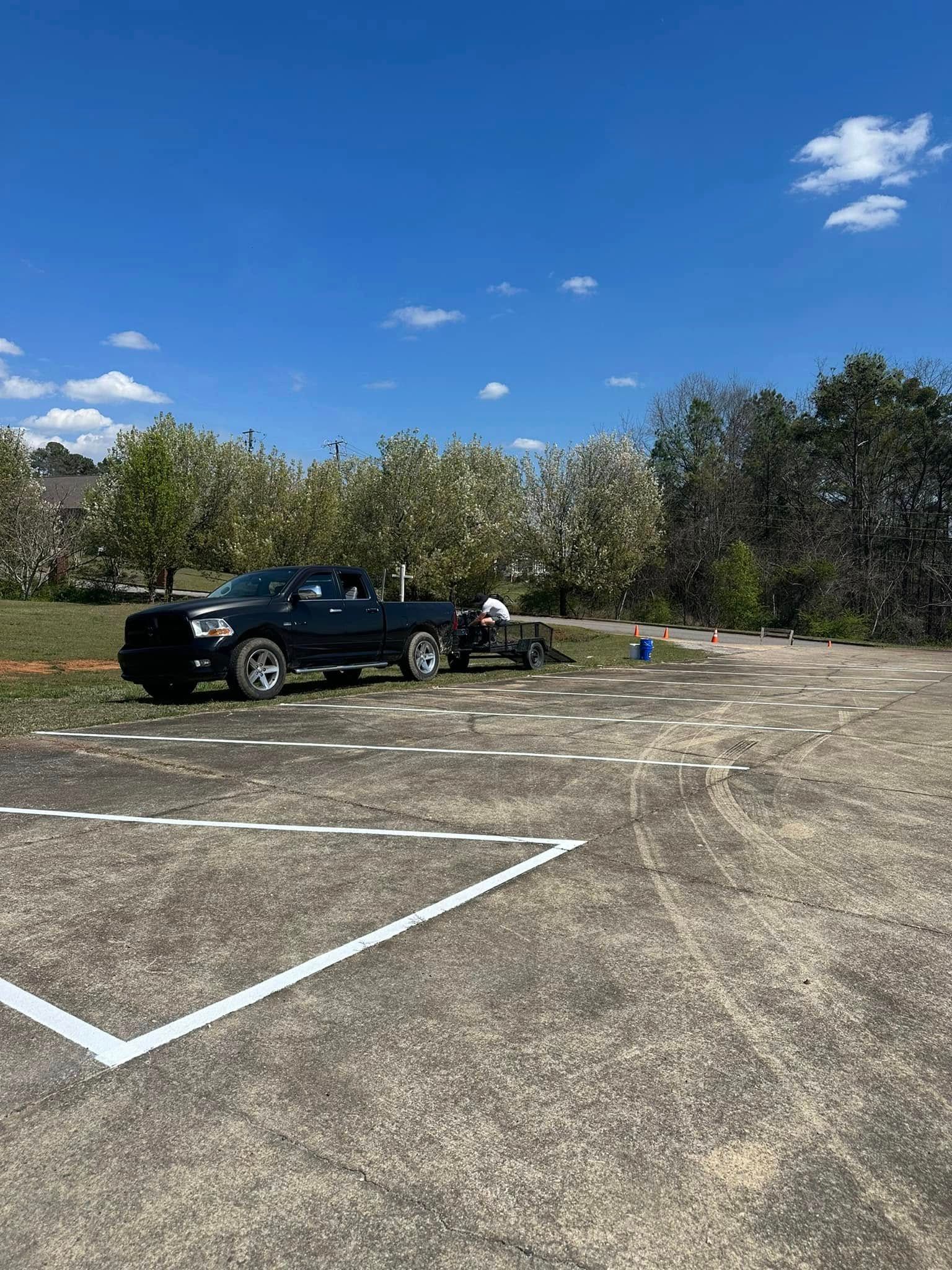 Black truck with trailer parked on dirt lot marked with white lines, blue sky, trees in background.
