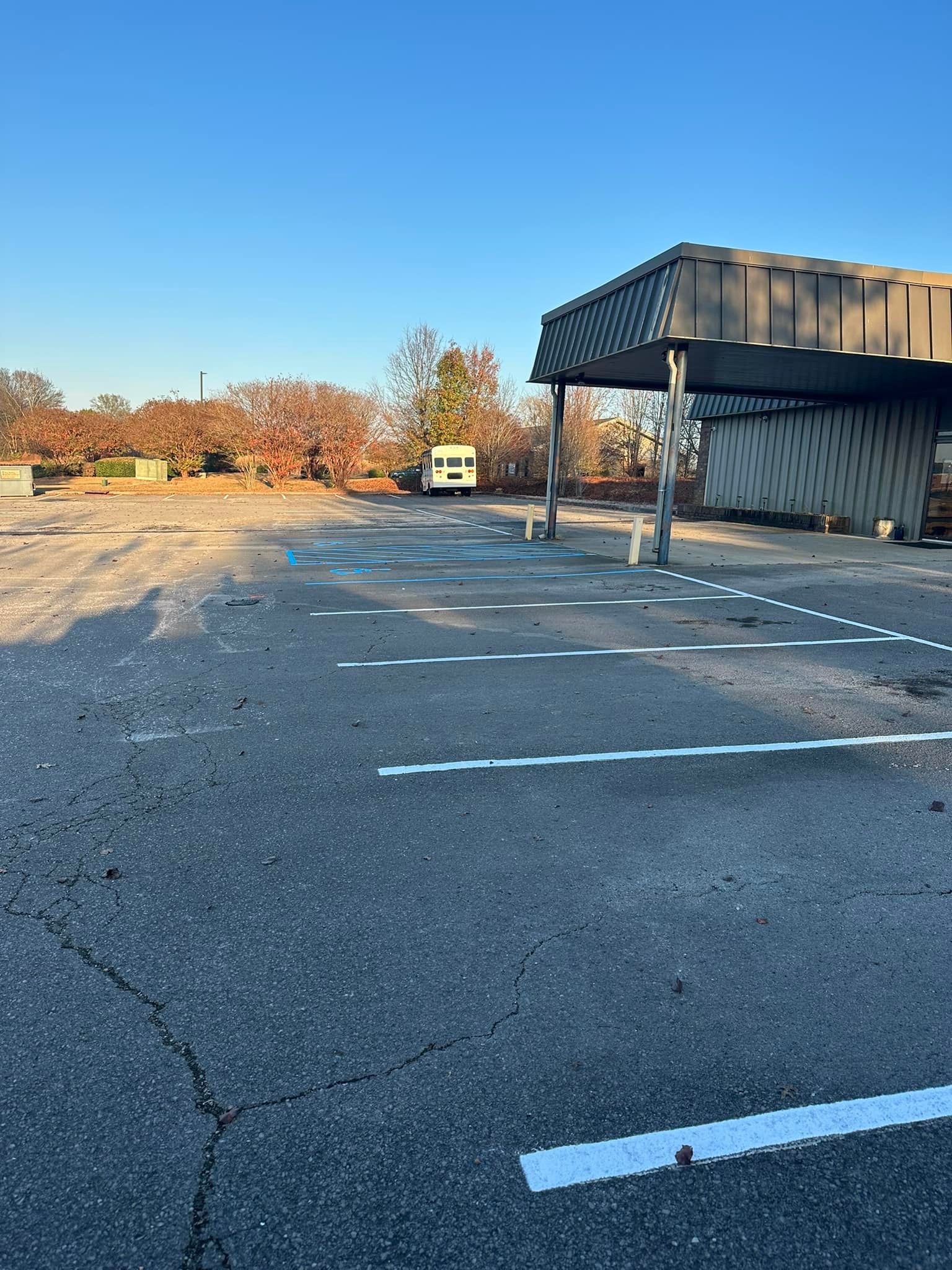 Empty parking lot with faded white lines, awning, trees, and clear blue sky.