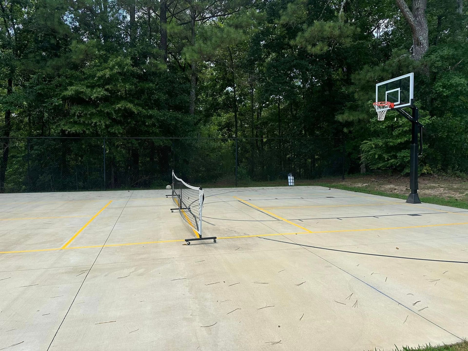 A concrete sports court with a tennis net and basketball hoop. Trees form a backdrop.
