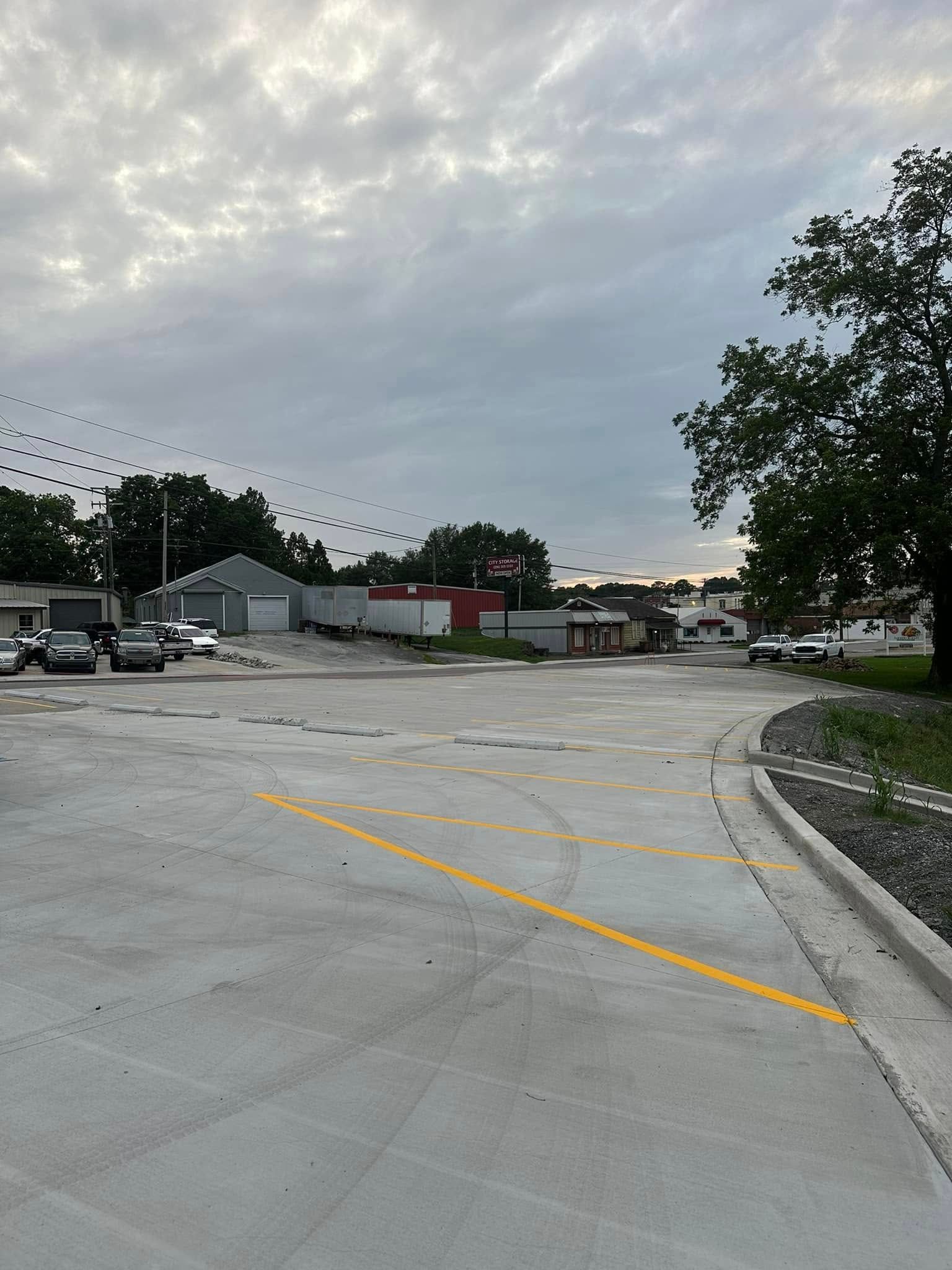 New concrete lot with yellow painted lines; buildings and trees in the background under a cloudy sky.