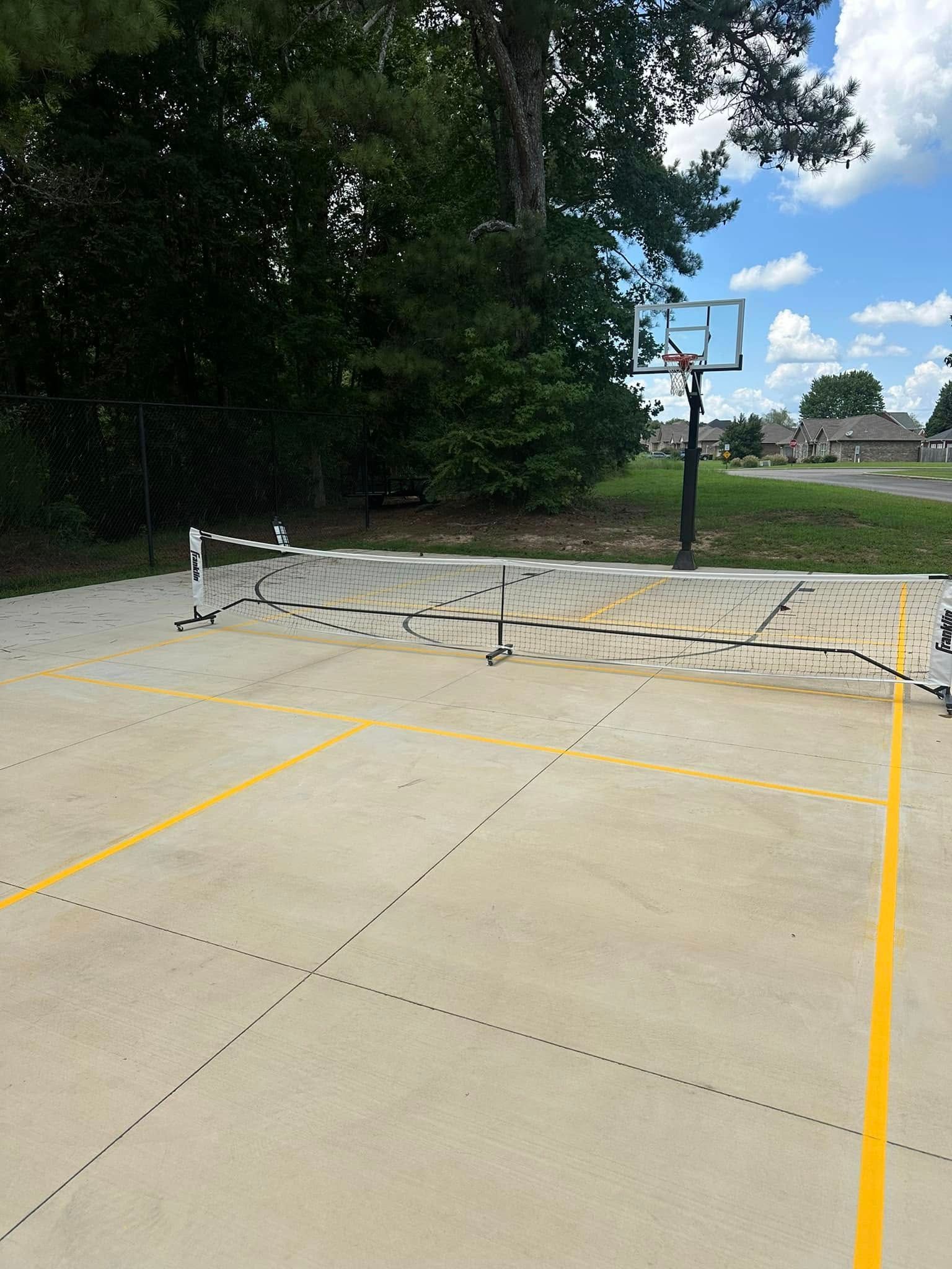 Pickleball court with net and yellow lines, basketball hoop in the background, trees, and a partly cloudy sky.