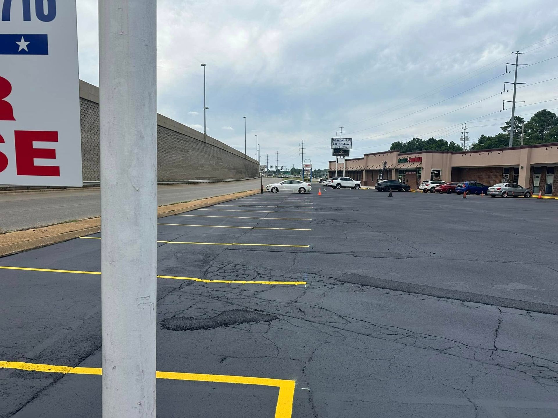 Large empty parking lot with yellow lines, a pole, and a row of shops on a cloudy day.