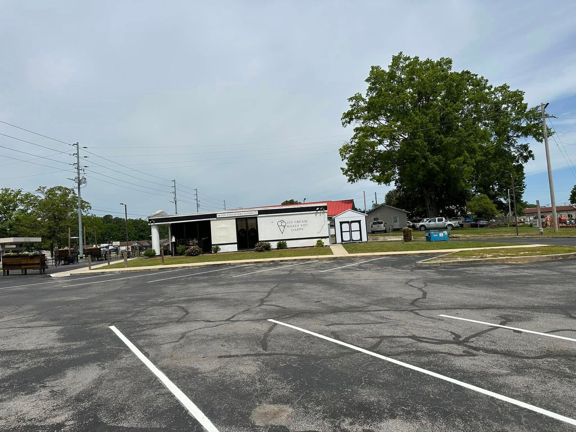 A small, white business with a red roof, located in a parking lot. A large tree is behind it under a cloudy sky.