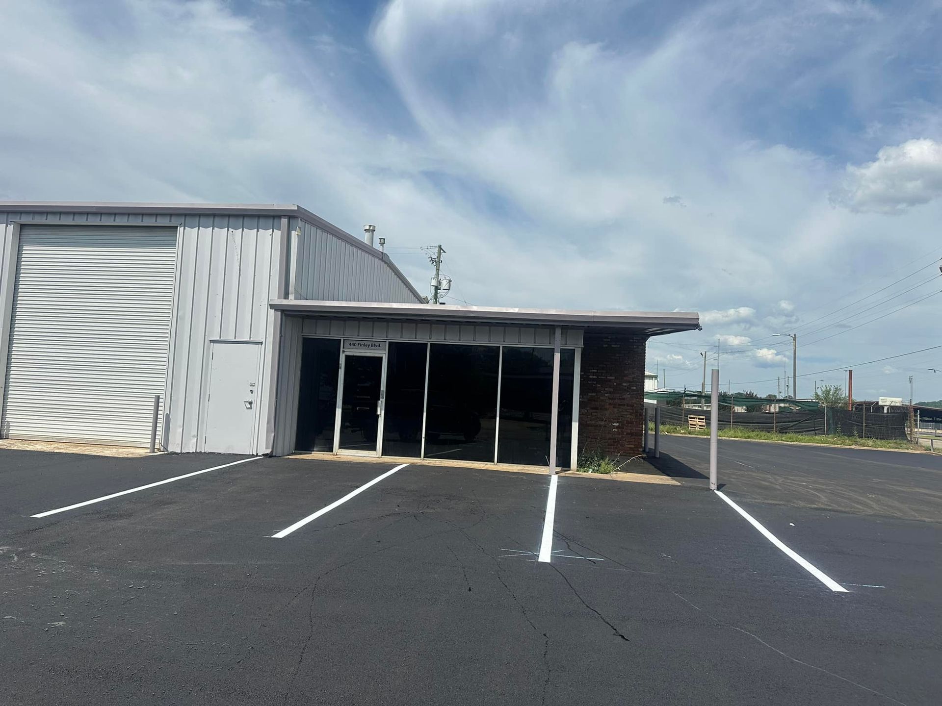 A gray metal building with a small storefront, set in a paved parking lot with white lines under a cloudy sky.