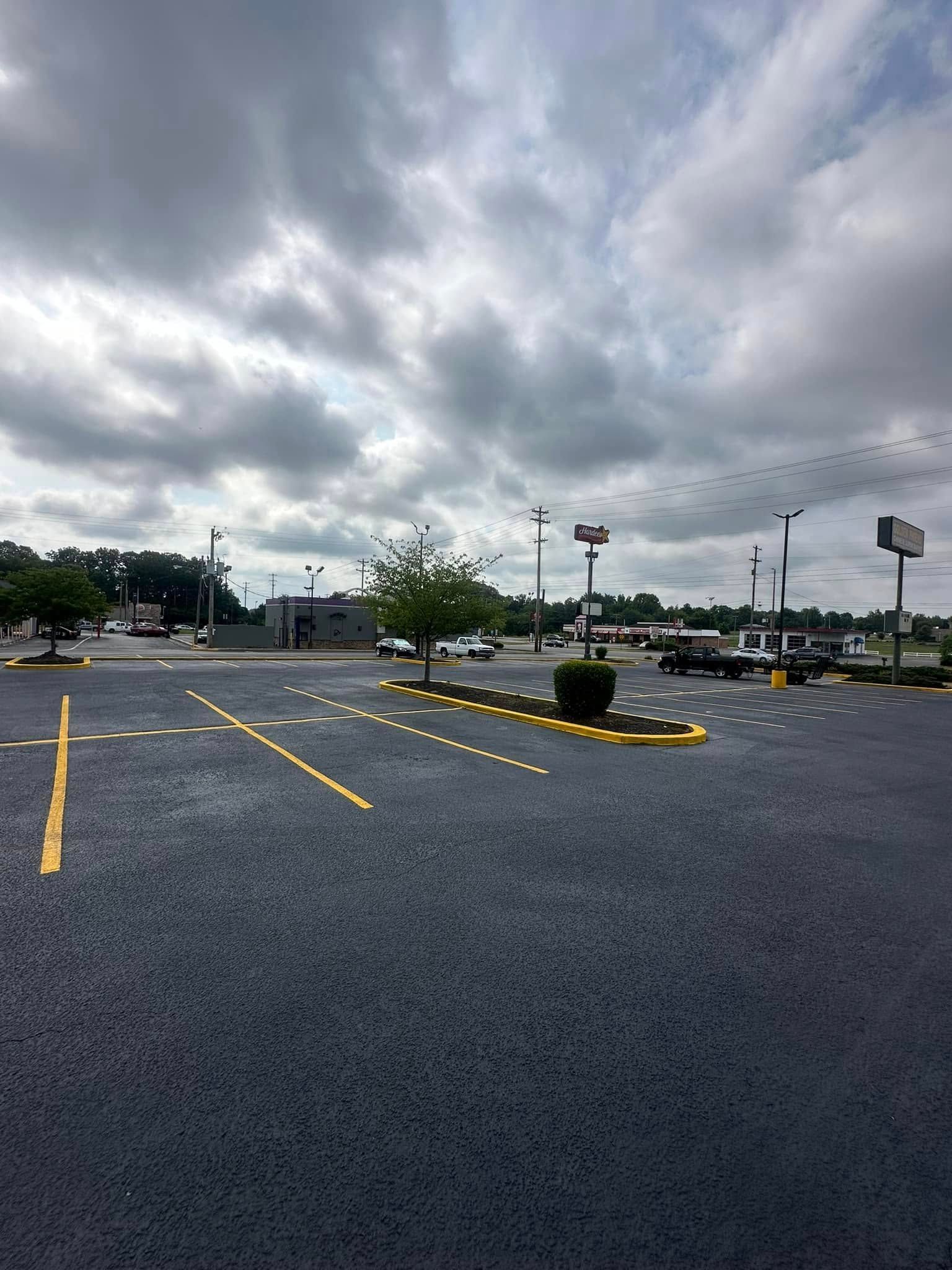 Empty asphalt parking lot under a cloudy sky. Yellow parking lines, trees, and distant buildings.