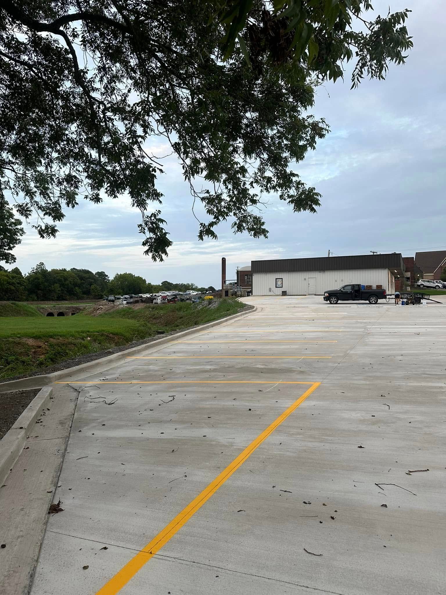 Empty, newly paved parking lot with yellow lines. White building and parked truck in the background, under cloudy sky.