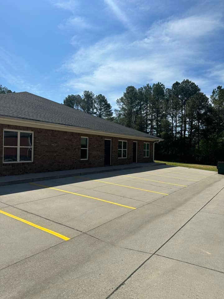 Exterior view of a brick building with a parking lot marked with yellow lines; trees in the background, blue sky.