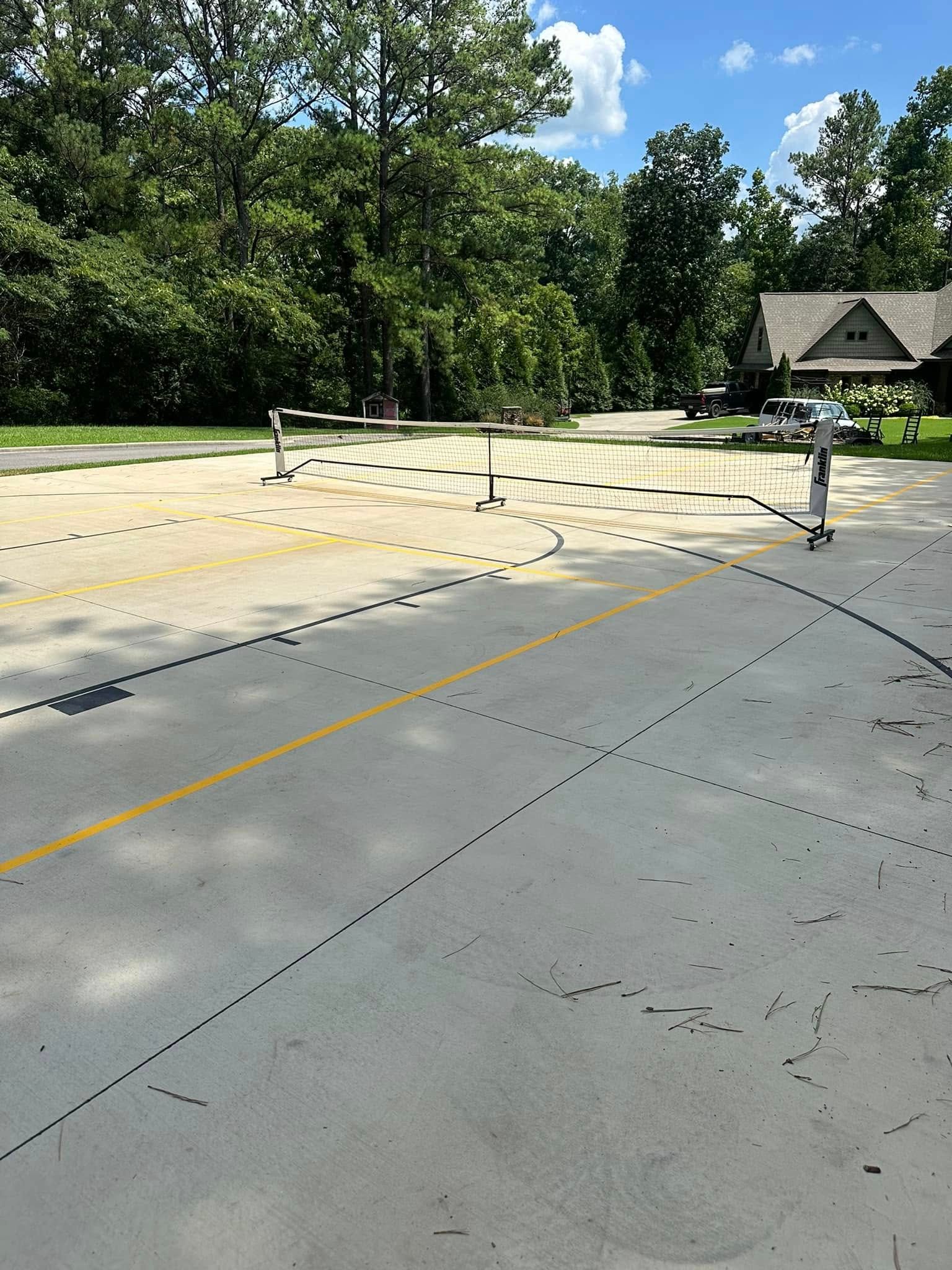 Paved area with dotted lines, yellow center line, and poles. Trees and a house are in the background.