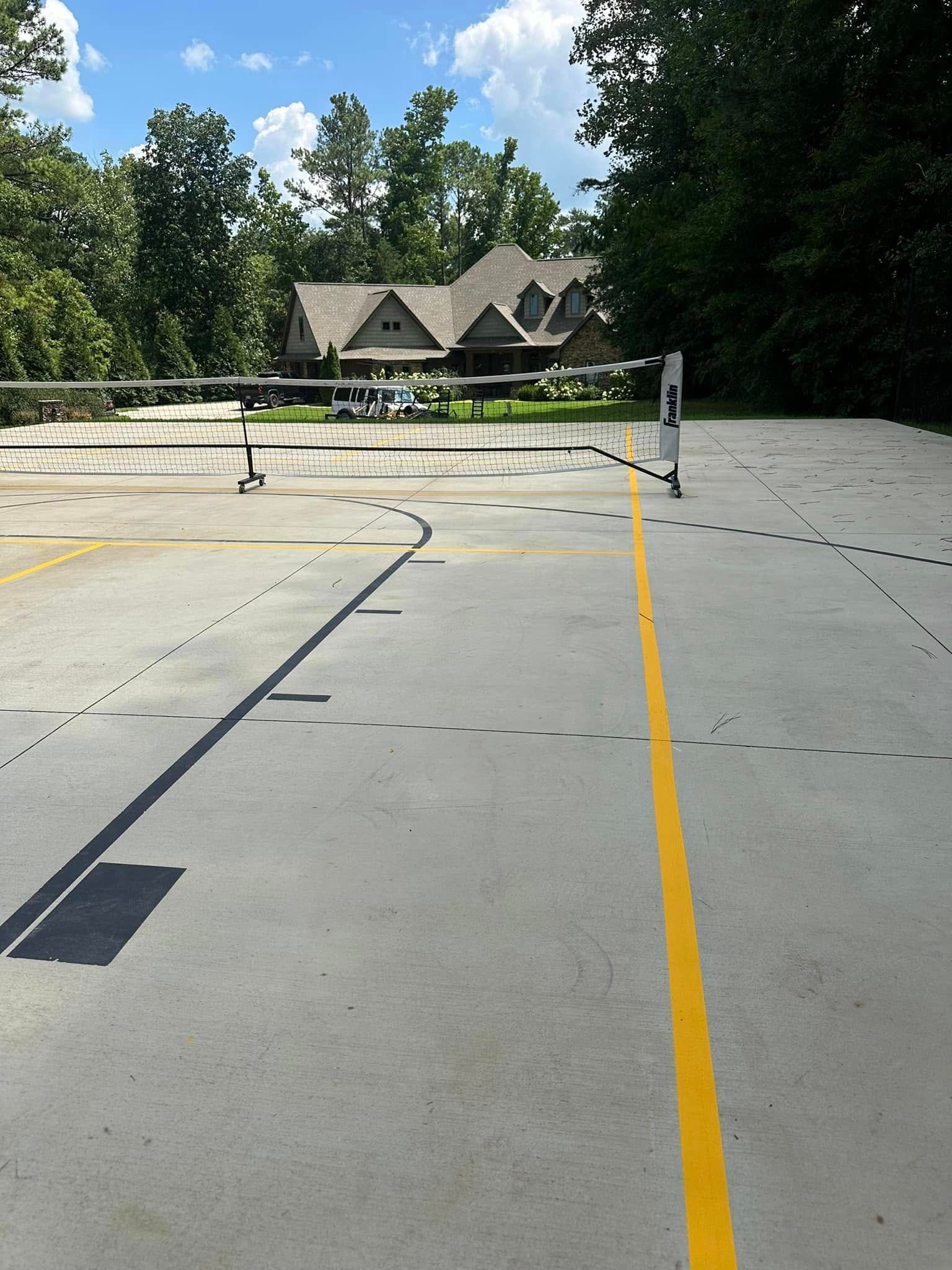 A tennis court with yellow lines, a net, and a house in the background. Sunny day.